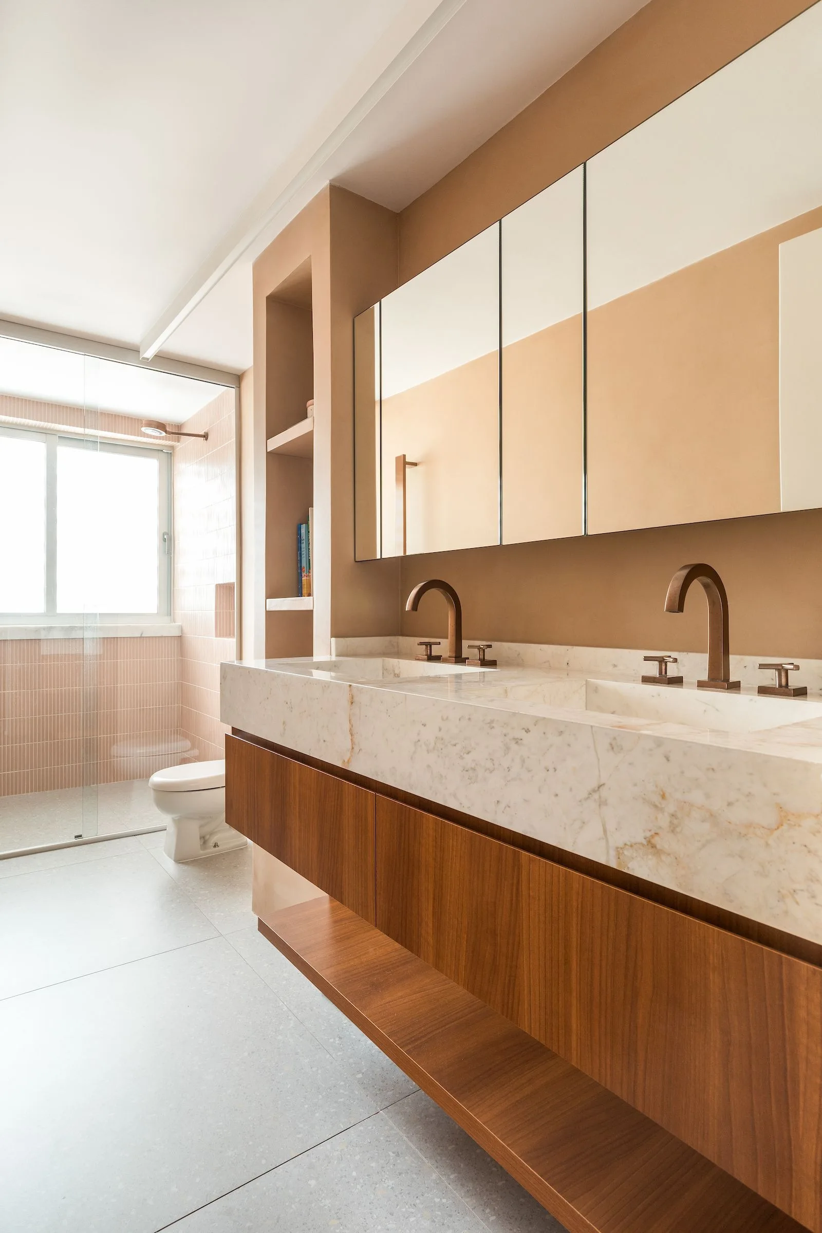 A contemporary bathroom features a floating wood vanity with a white marble countertop, bronze faucets, and a mirrored medicine cabinet against tan walls.