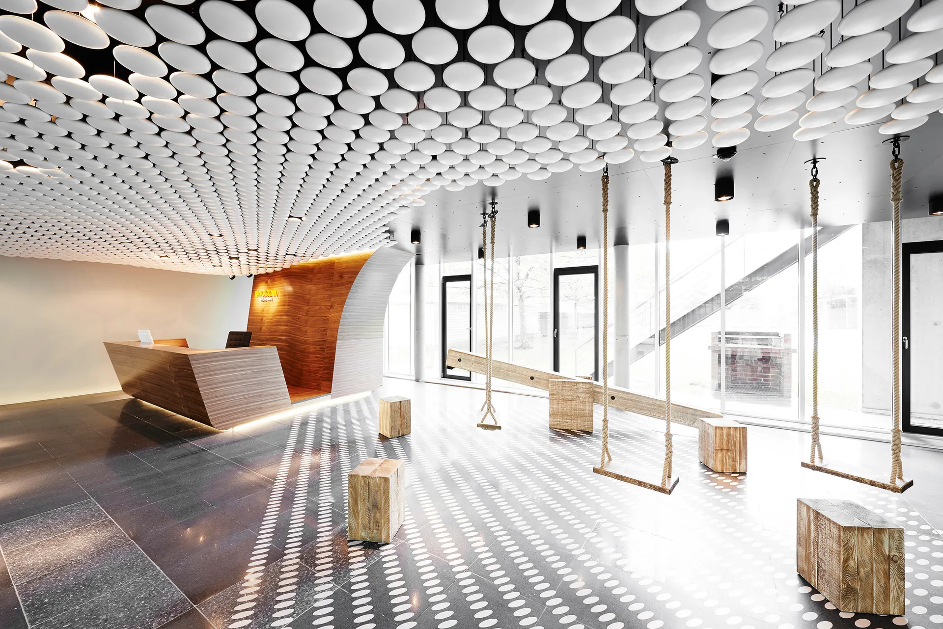 A modern lobby features a ceiling of white circular discs, a wooden reception desk, and rope swings suspended over a patterned grey tile floor.