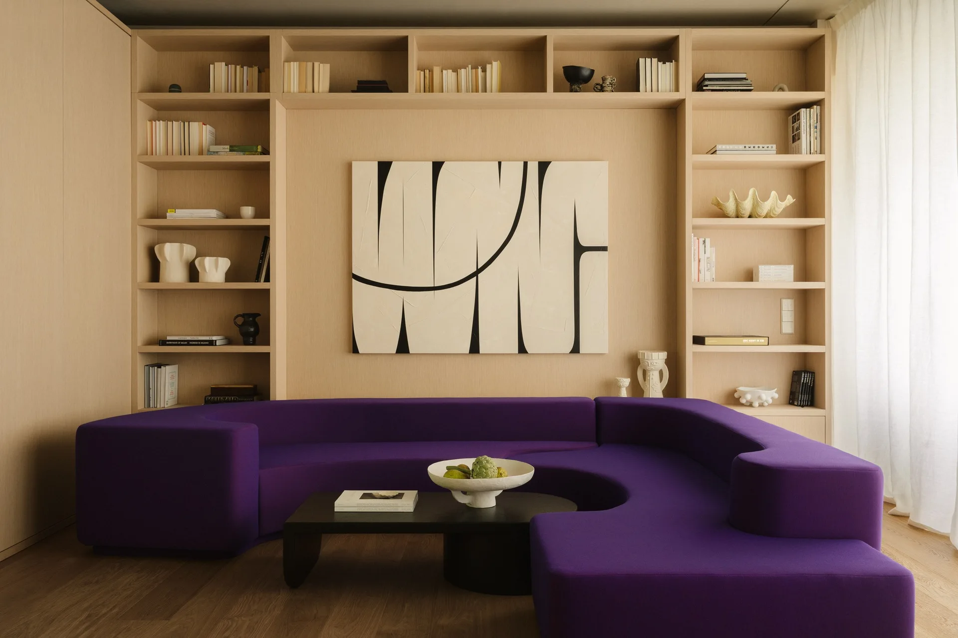 A vibrant purple curved sofa is positioned in front of a symmetrical light wood built-in shelving unit holding books and a large abstract painting.