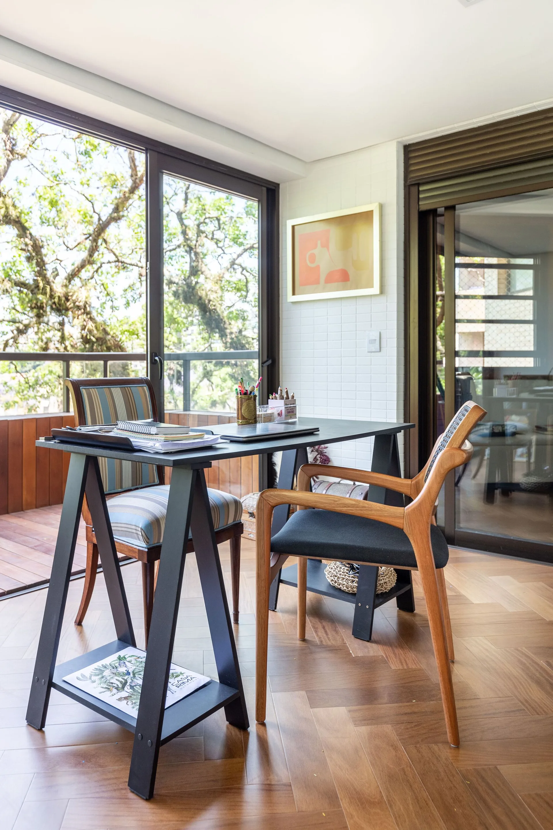 A home office features a black trestle desk with two mismatched chairs on a rich herringbone wood floor next to large sliding glass doors.