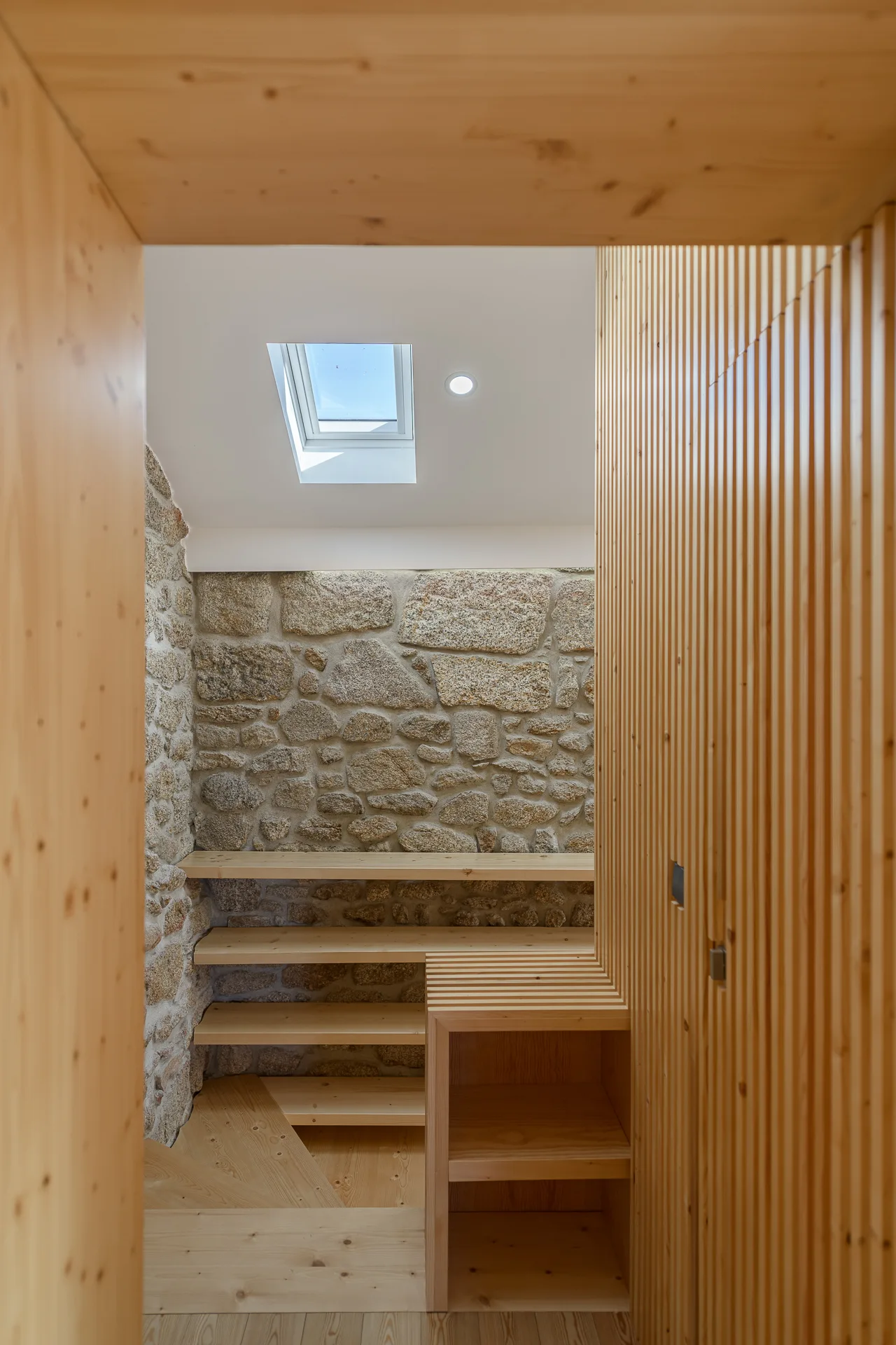 A light wood staircase and built-in shelving are set against a rough stone wall, lit by a ceiling skylight.