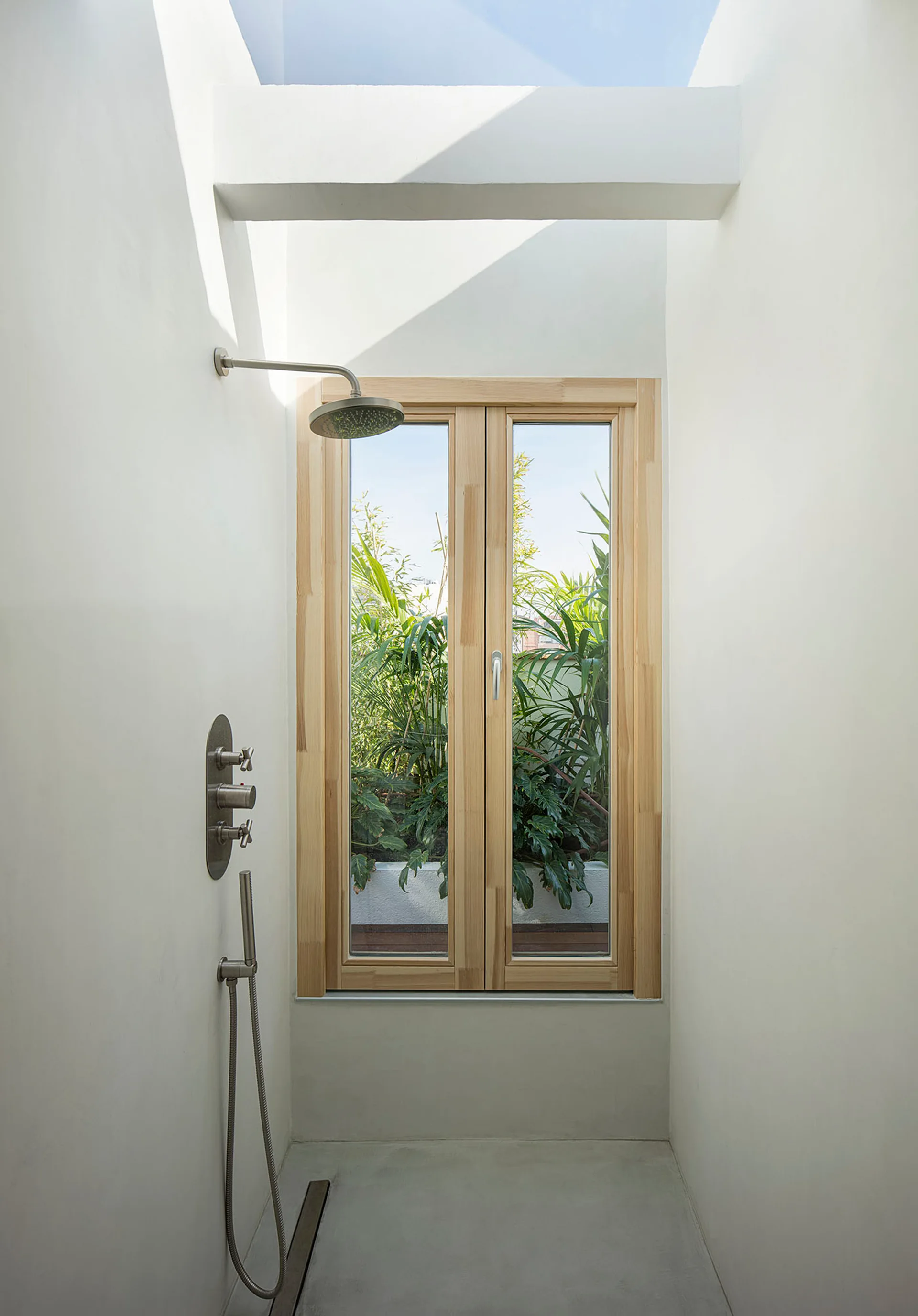 A white plaster shower stall with a light wood window overlooking greenery, stainless steel fixtures, and an overhead skylight.