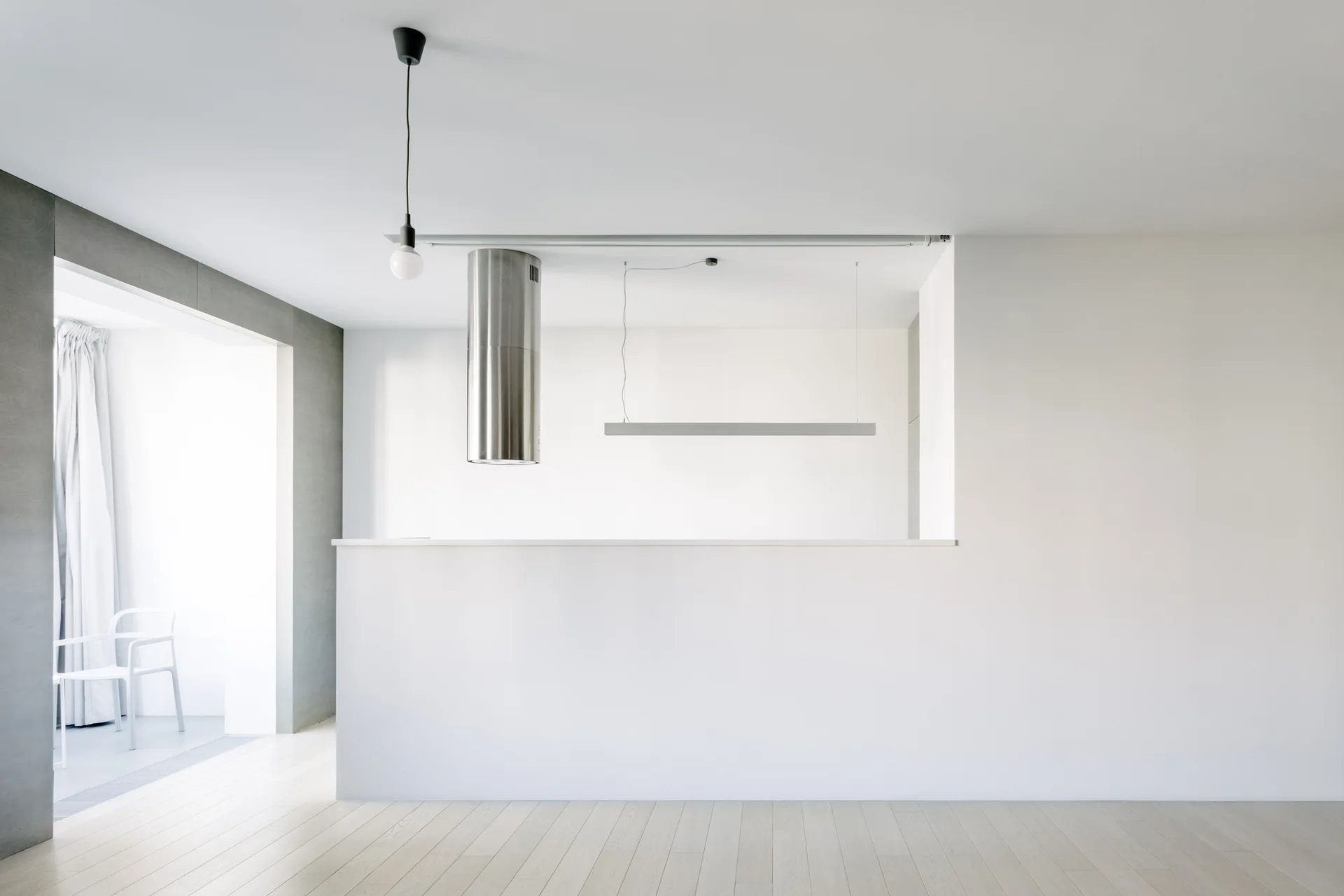 A minimalist white kitchen features a partition wall, a cylindrical stainless steel range hood, and a slim pendant light over pale wood floors.