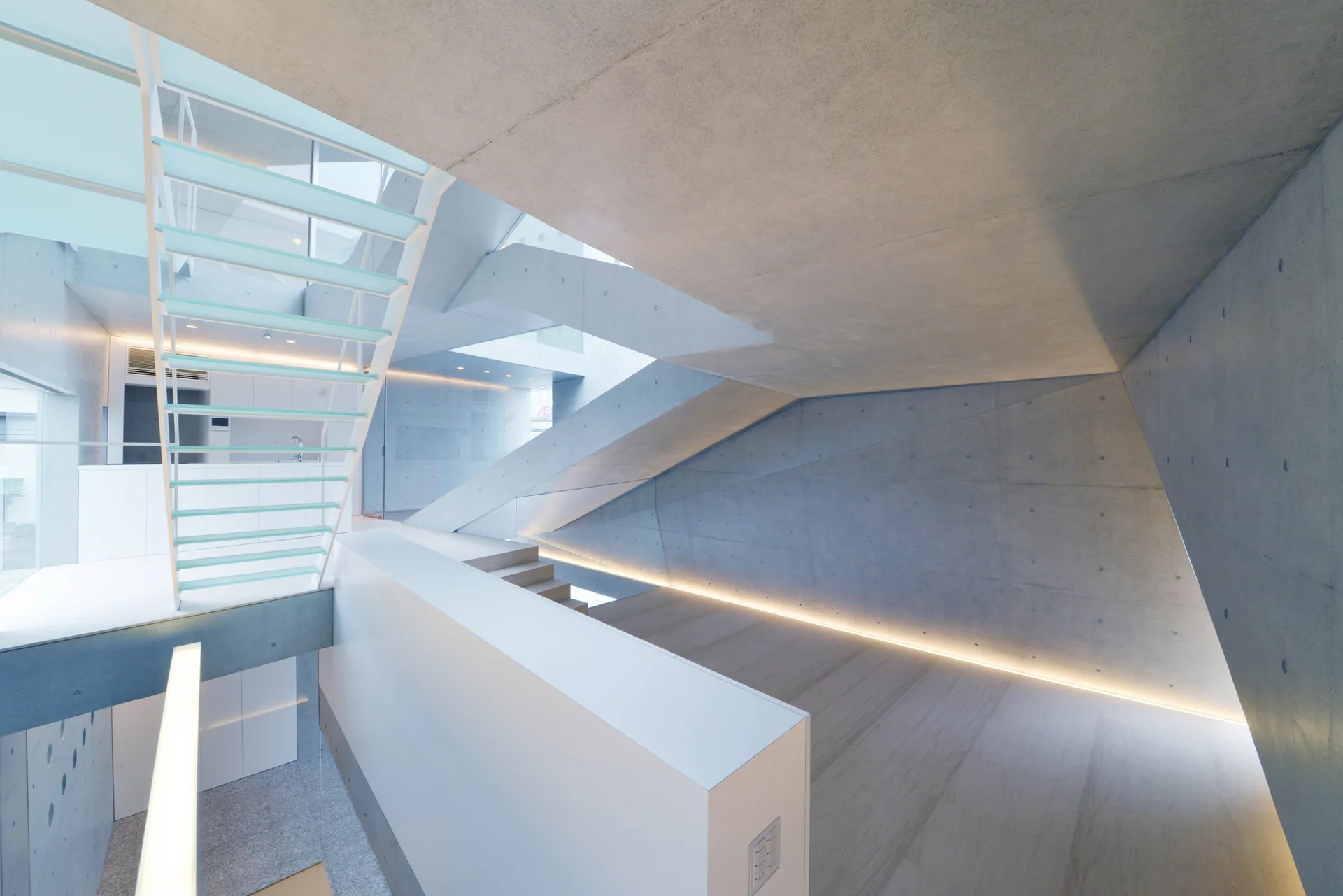 A minimalist multi-level concrete staircase features floating glass treads, white metal railings, and warm linear lighting along angled walls.
