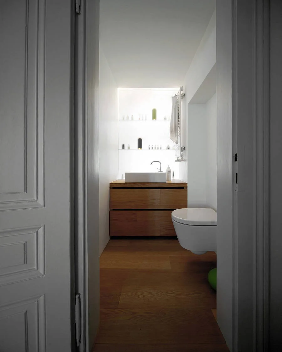 A view through a doorway into a narrow white bathroom featuring a wooden vanity, a wall-mounted toilet, and backlit recessed shelving.