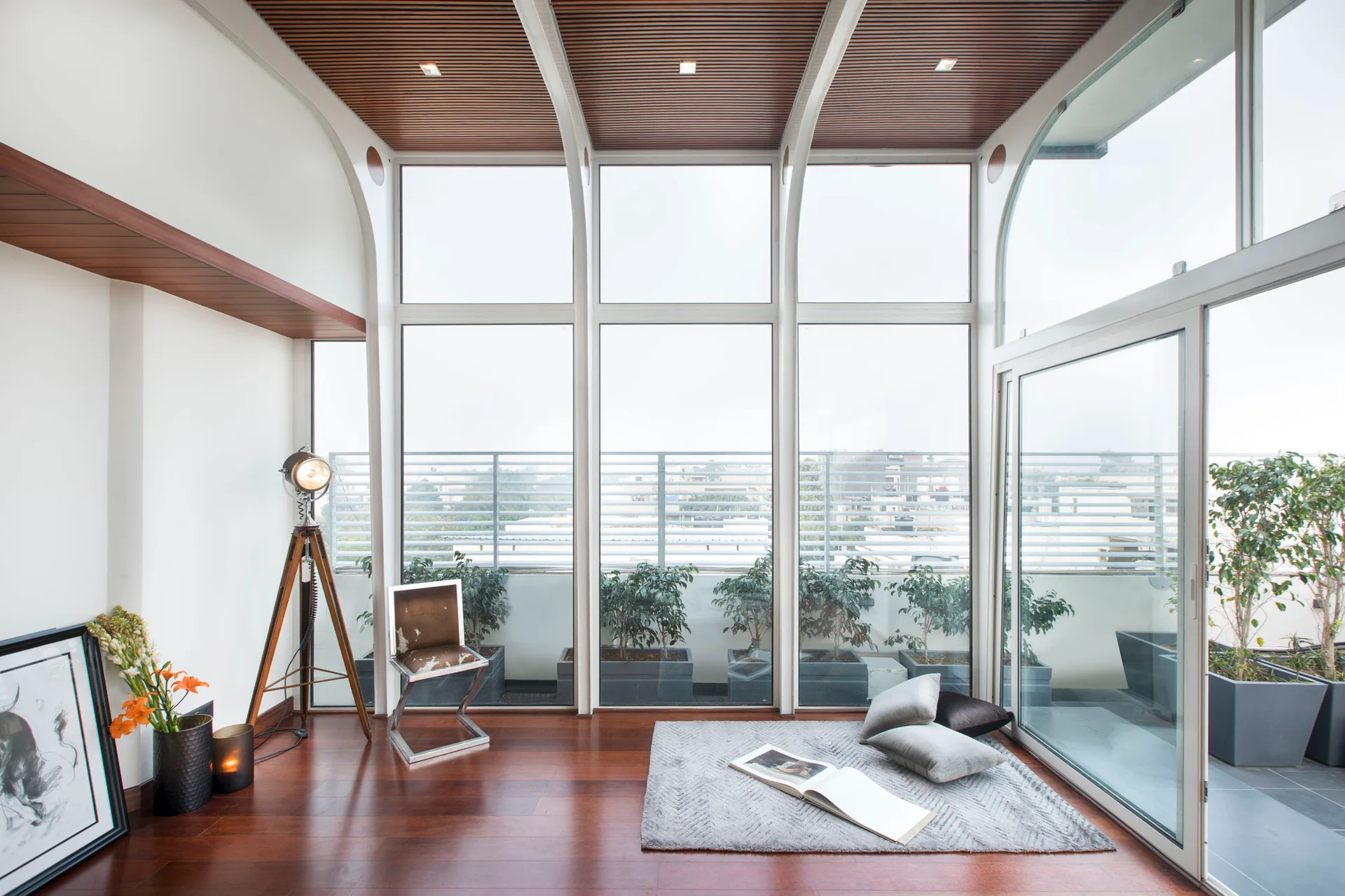 A modern sunroom features a slatted wood ceiling, arched support beams, floor-to-ceiling windows, and warm hardwood floors with a small grey rug.