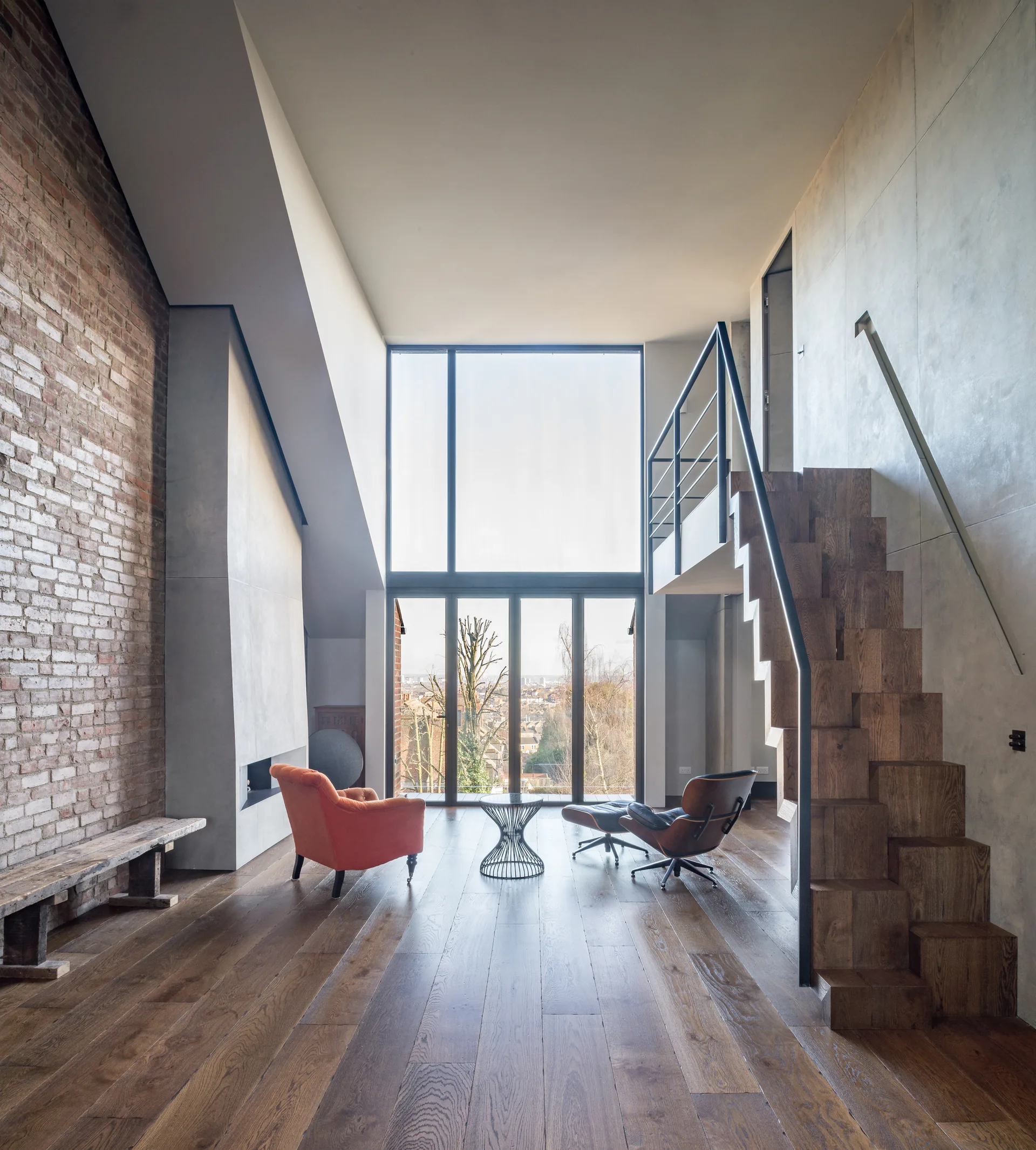 A tall living room featuring a geometric wooden block staircase, an exposed brick wall, and two modern lounge chairs facing a large two-story window.