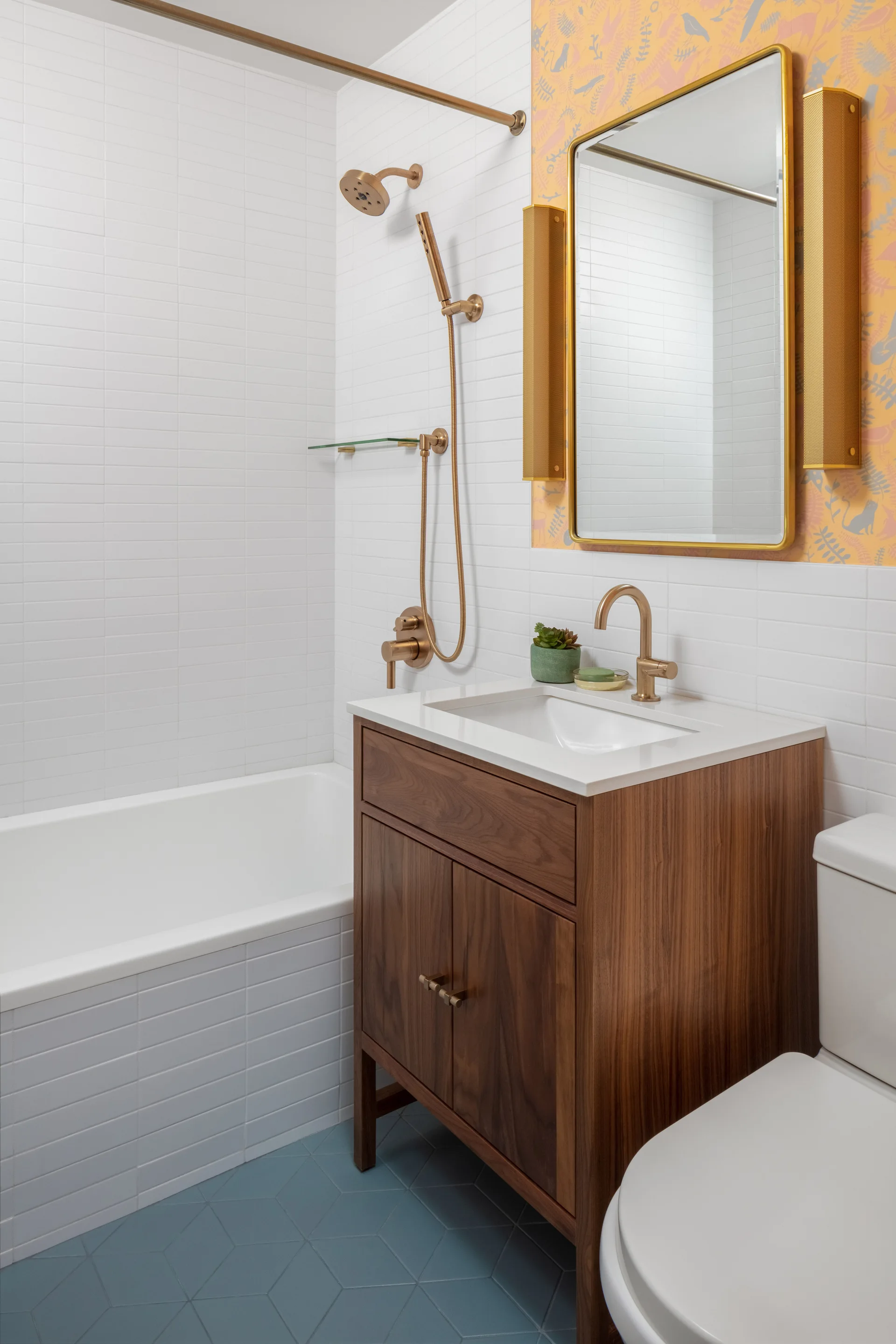 A compact bathroom features a wood vanity, white tiled walls, blue geometric floor tiles, and yellow patterned wallpaper behind a gold-framed mirror.