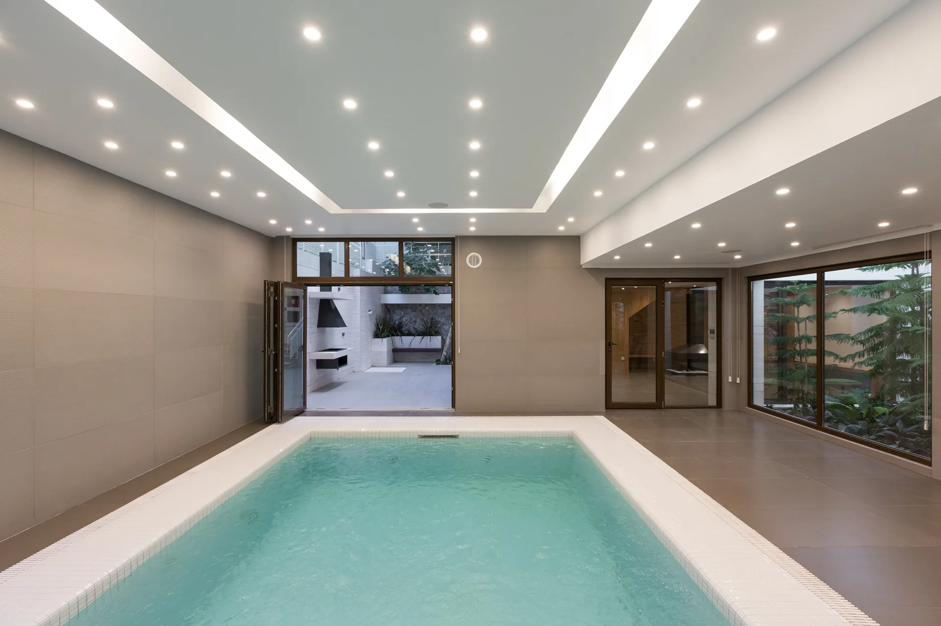 An indoor swimming pool features a white tile rim, grey walls, and a large tray ceiling illuminated by multiple small recessed lights.