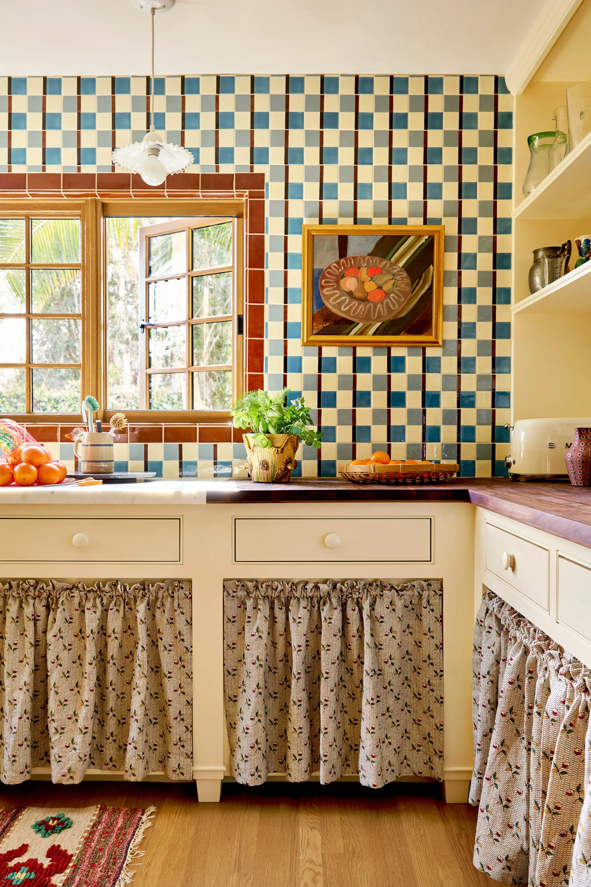 A rustic kitchen features blue and cream checkerboard wall tiles, cream cabinets with floral print fabric skirts, and a wooden grid window.