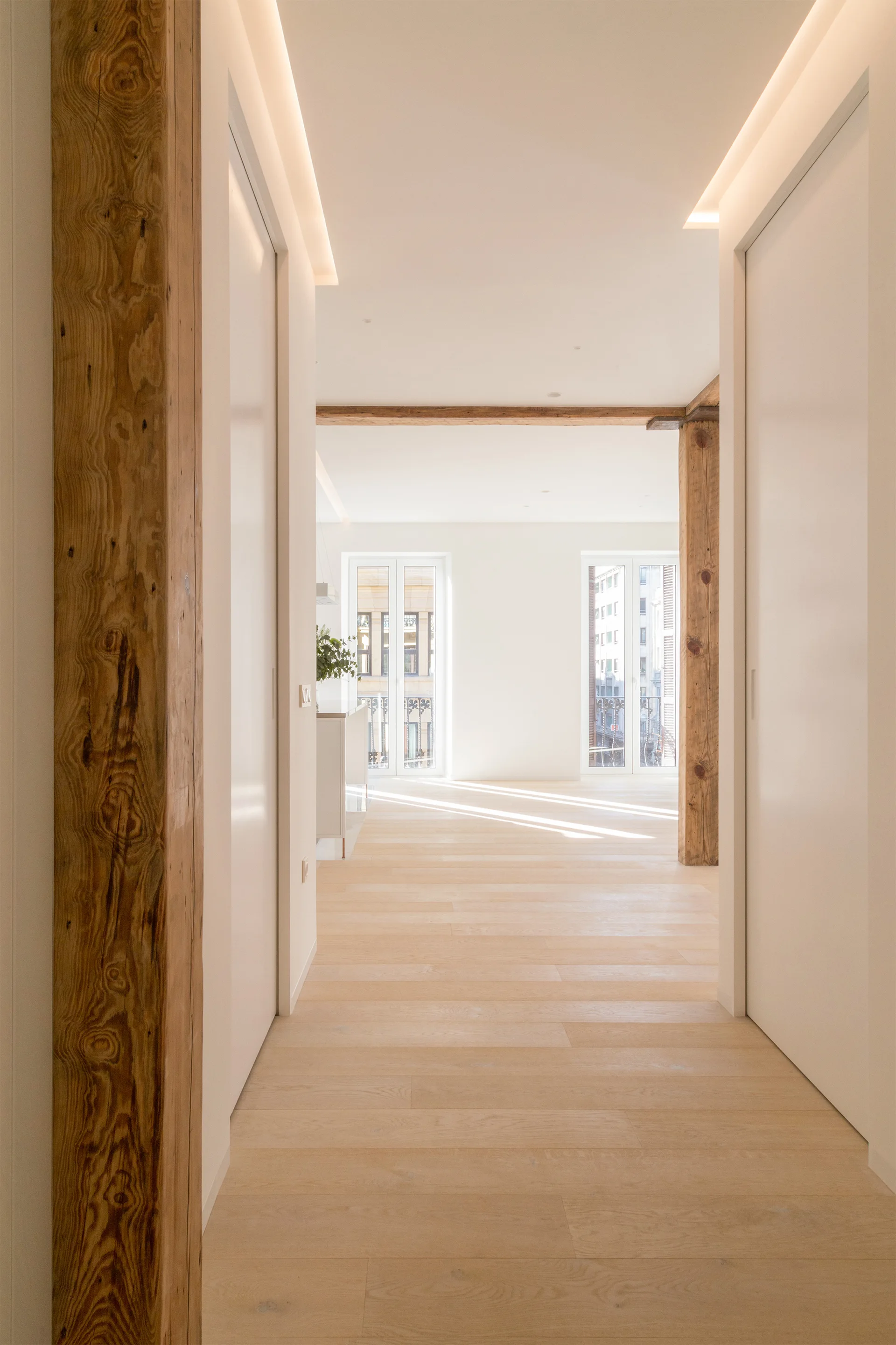 A bright minimalist hallway featuring light wood floors, smooth white walls with integrated doors, and rustic exposed wooden support columns.