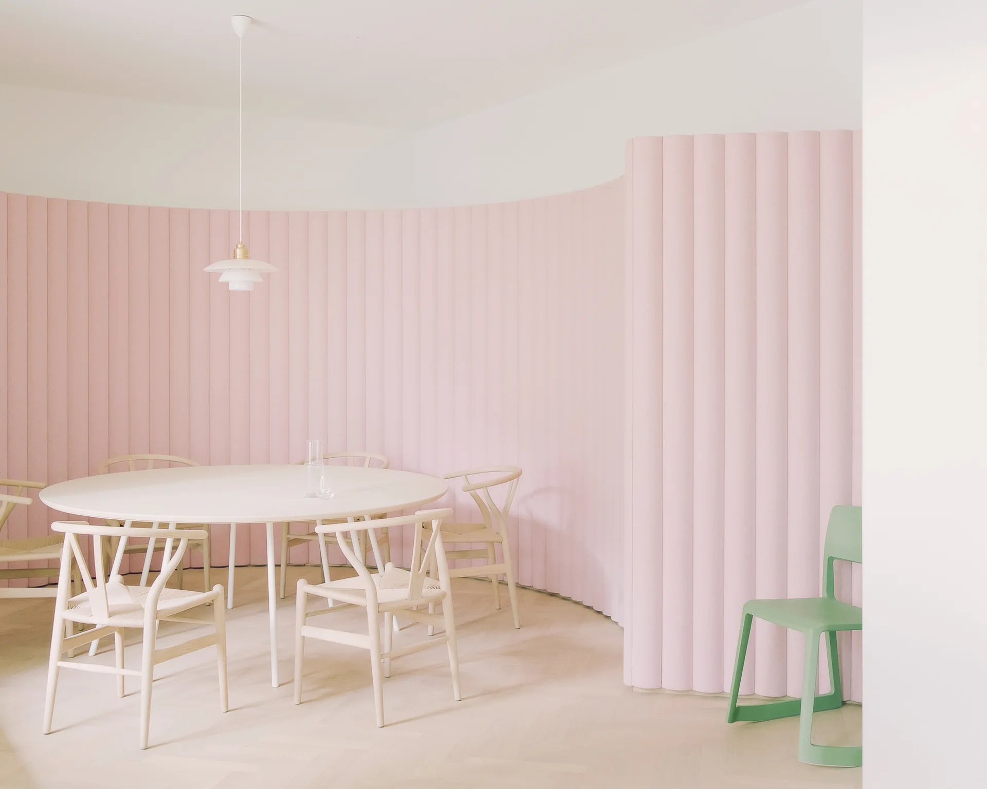 A white circular dining table with light wood wishbone chairs is set against a tall, curved, pale pink fluted wall on a herringbone floor.