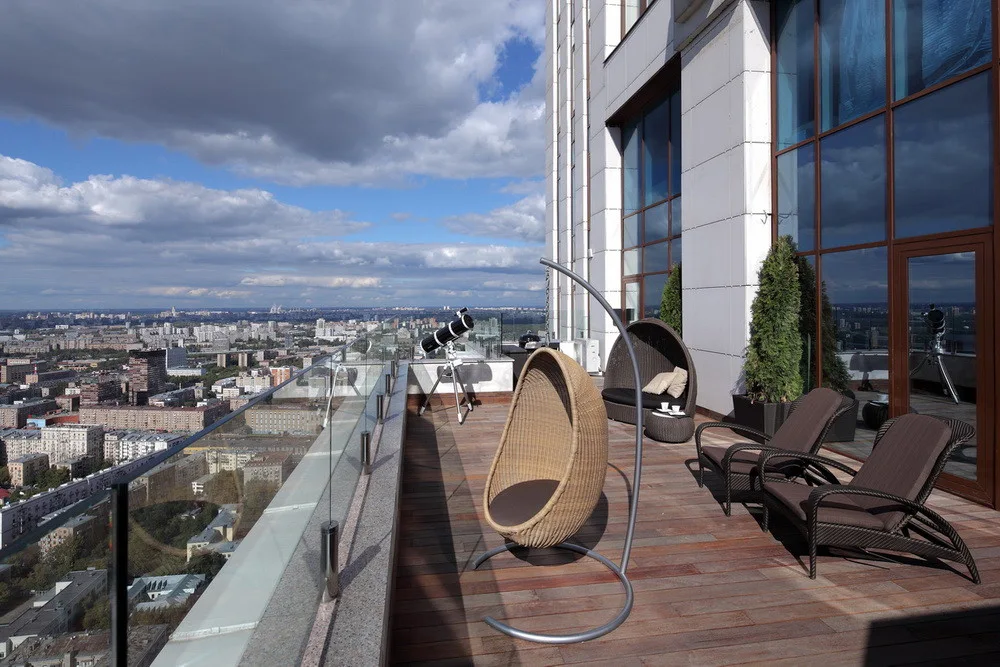 A spacious terrace features a wood plank deck, a glass railing overlooking the city, a hanging wicker egg chair, and brown lounge seating.