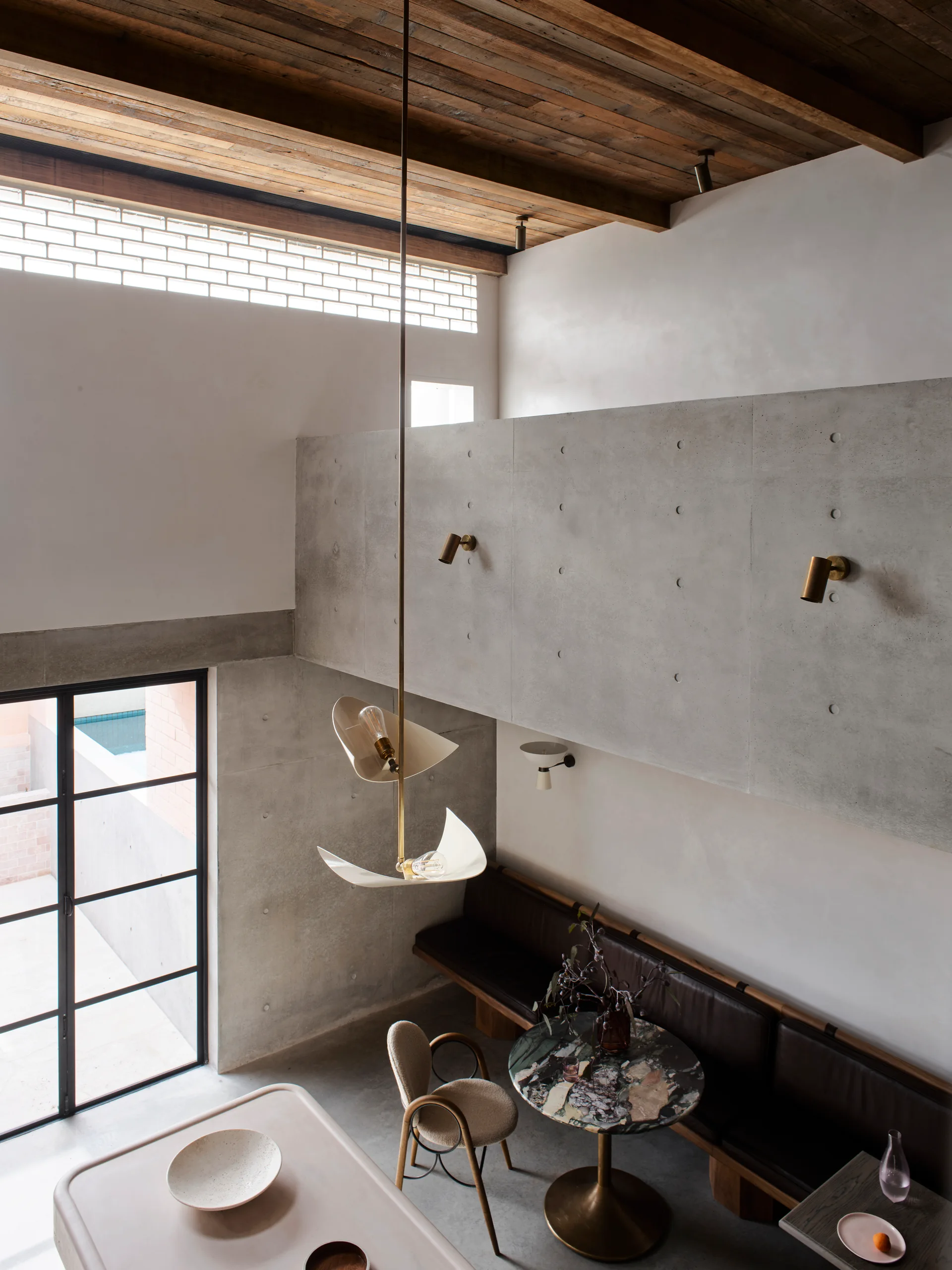 A high-angle view of a modern dining room featuring a round marble table, a built-in leather bench, concrete walls, and an exposed wood ceiling.