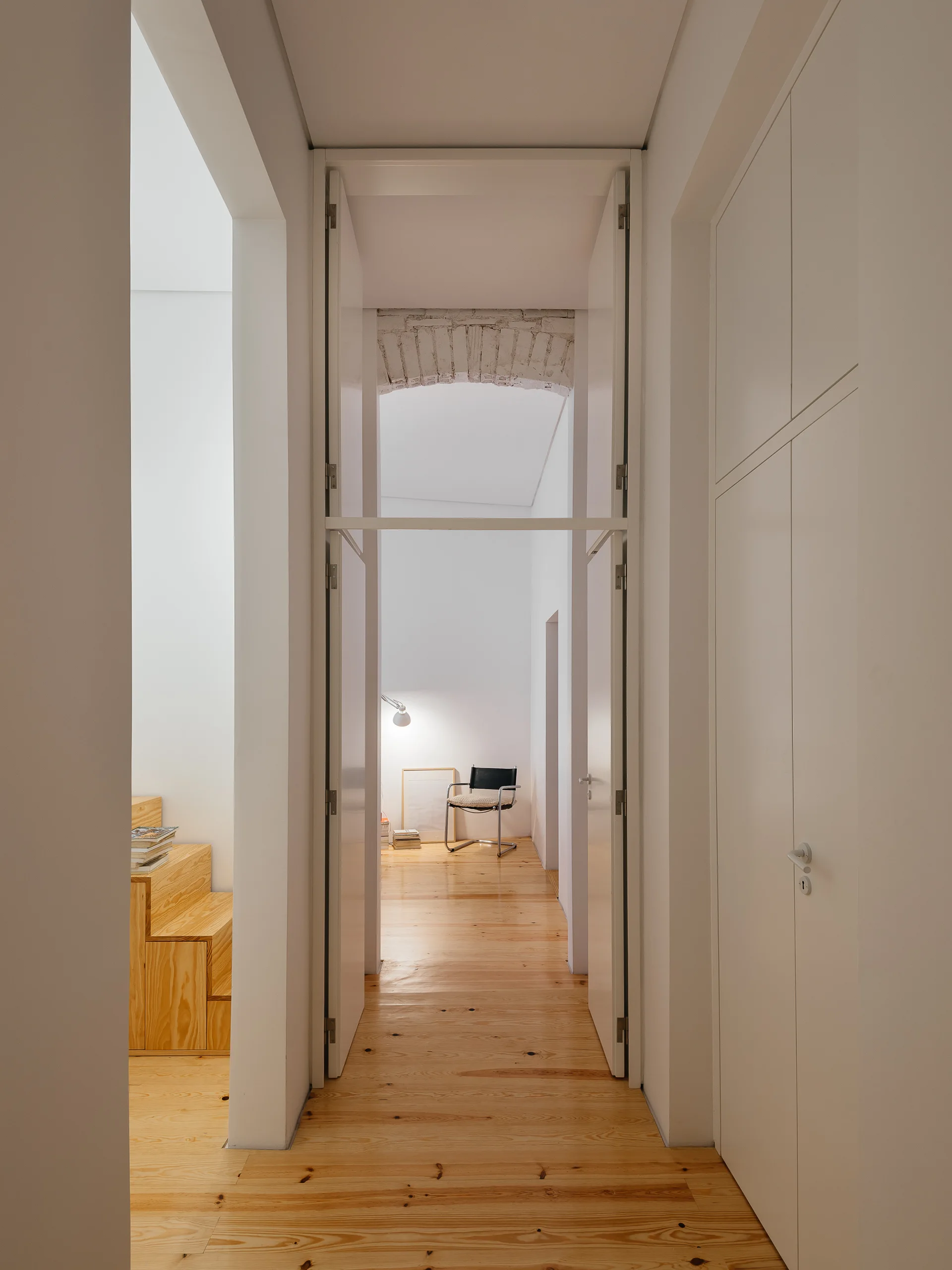 A minimalist white hallway features light wood flooring, floor-to-ceiling built-in storage, and a view of a room with a metal-framed chair.