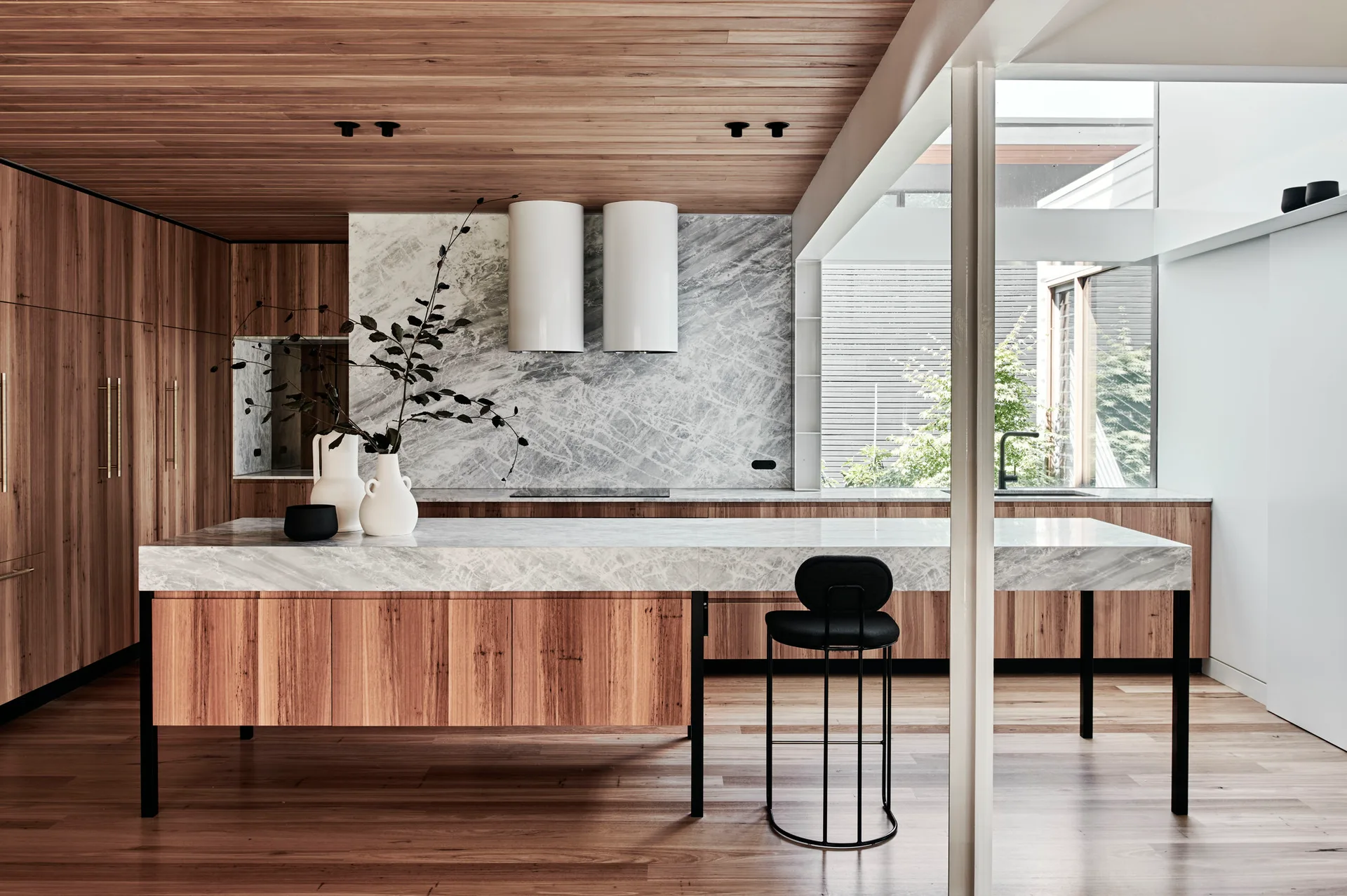 A modern kitchen features a long marble-topped island with wood cabinets, a matching wood slat ceiling, and two white cylindrical range hoods.