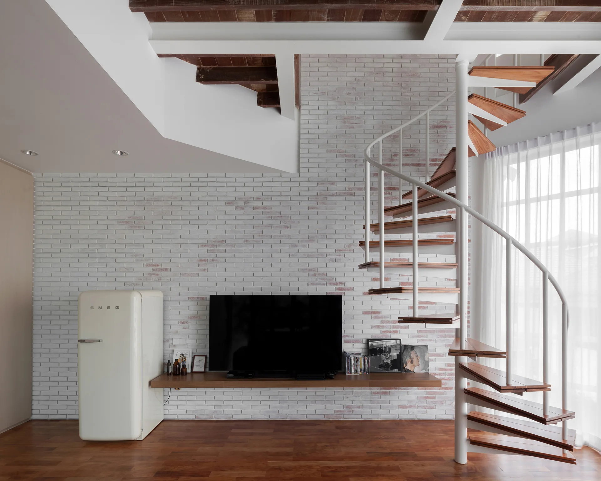 A white spiral staircase with wood treads sits in a living room featuring a whitewashed brick wall, a floating wooden shelf, and a cream refrigerator.