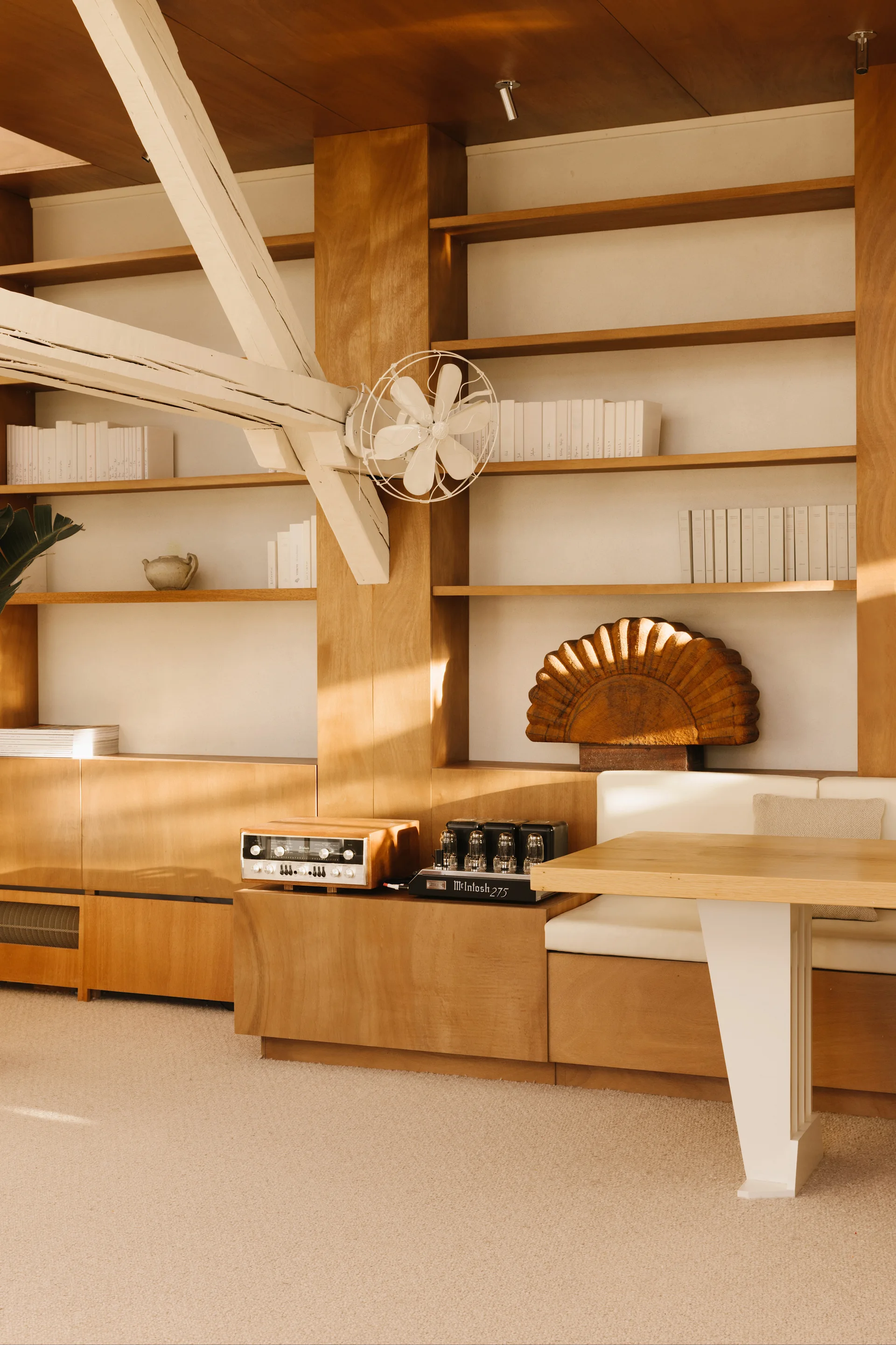 A wood-paneled room features floor-to-ceiling shelving, a built-in banquette with white cushions, a matching table, and a prominent white ceiling beam.