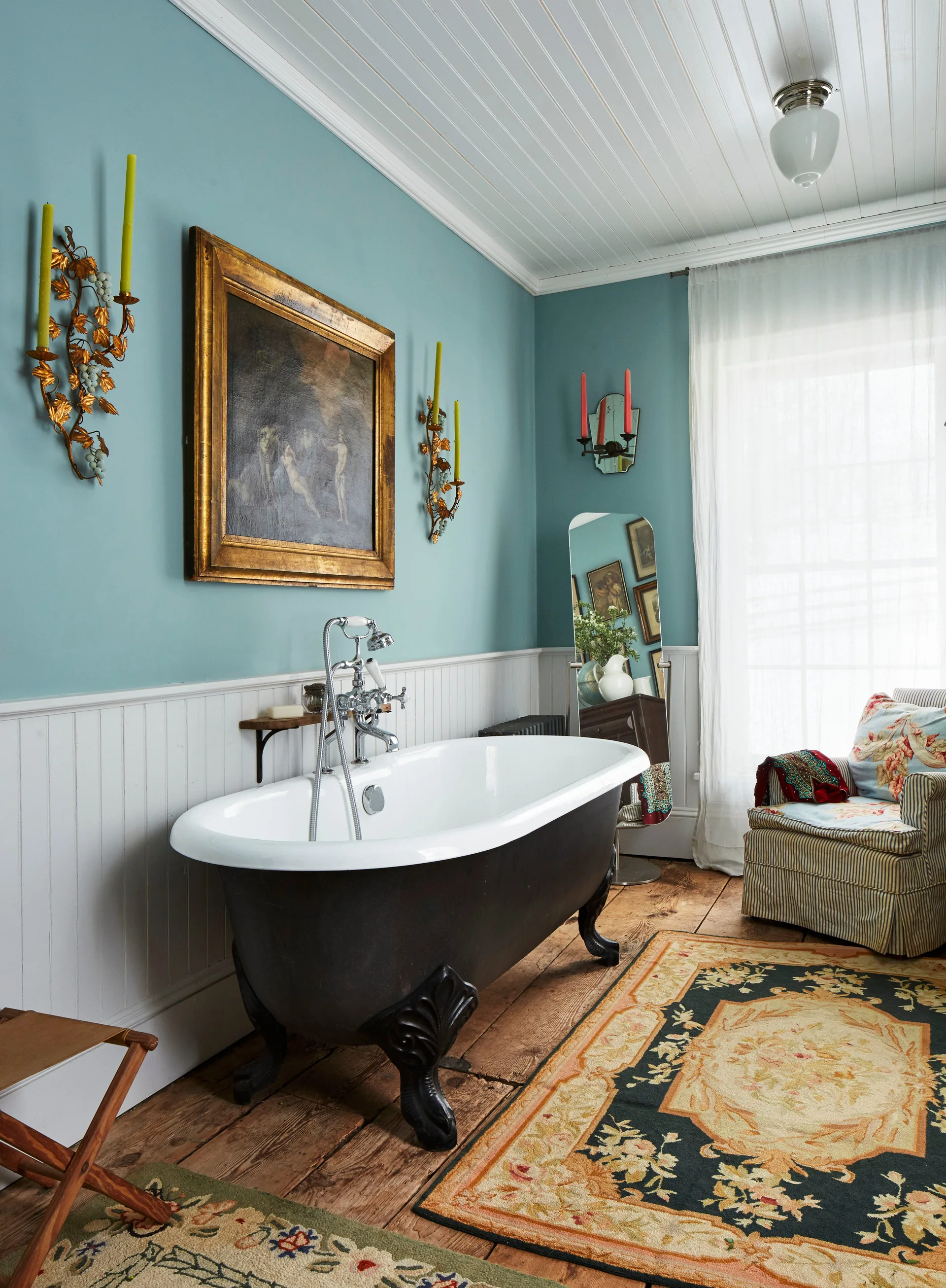 A black clawfoot bathtub sits on wood floors against teal walls with white beadboard wainscoting and a framed oil painting.