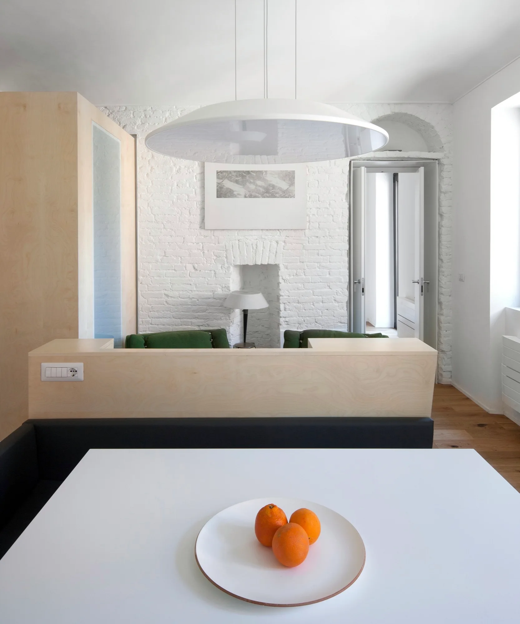 A white dining table with oranges sits before a light wood partition and a painted brick wall with an inset fireplace.