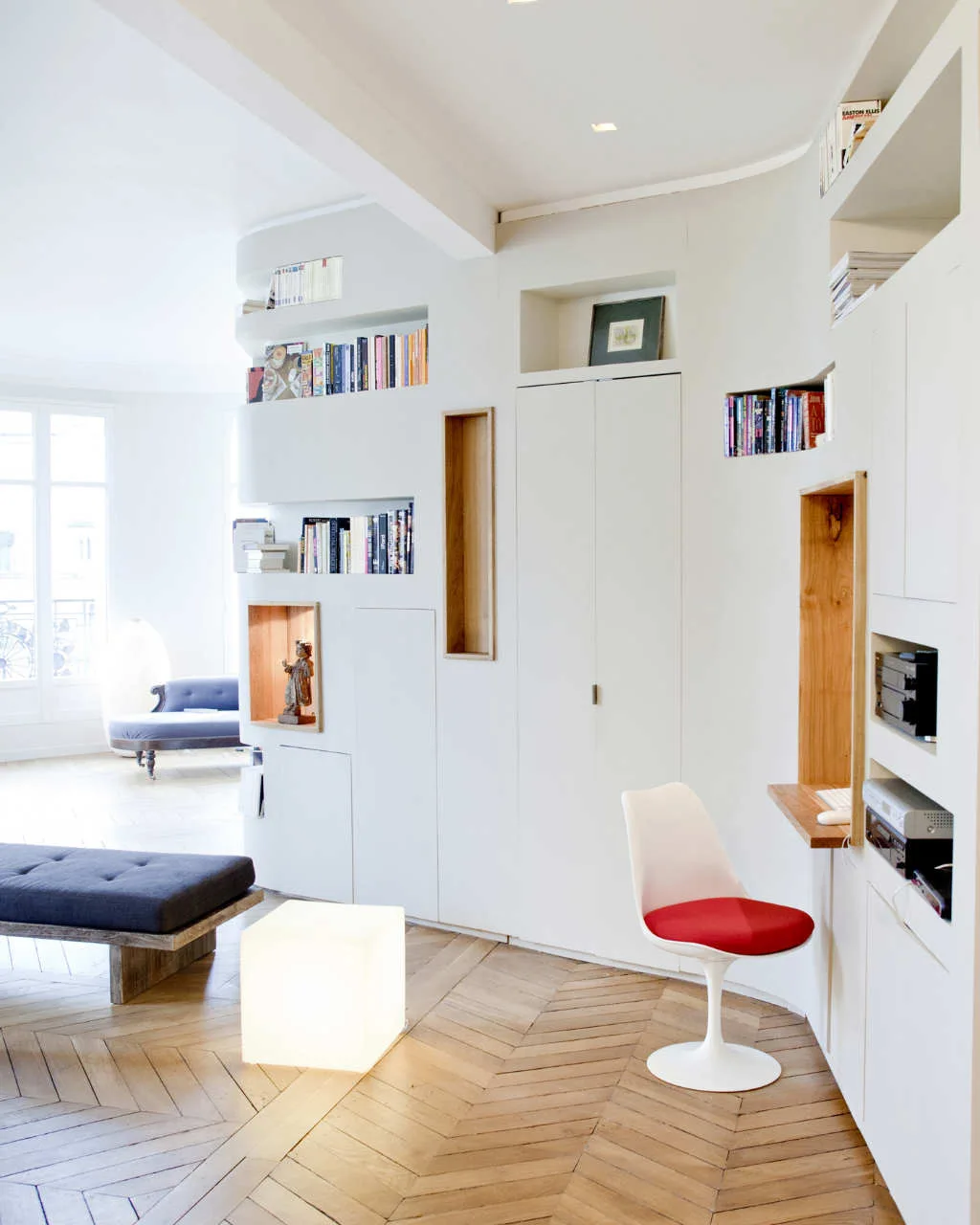 A custom curved white storage wall features recessed wood niches, an integrated desk with a red cushion chair, and herringbone parquet wood flooring.
