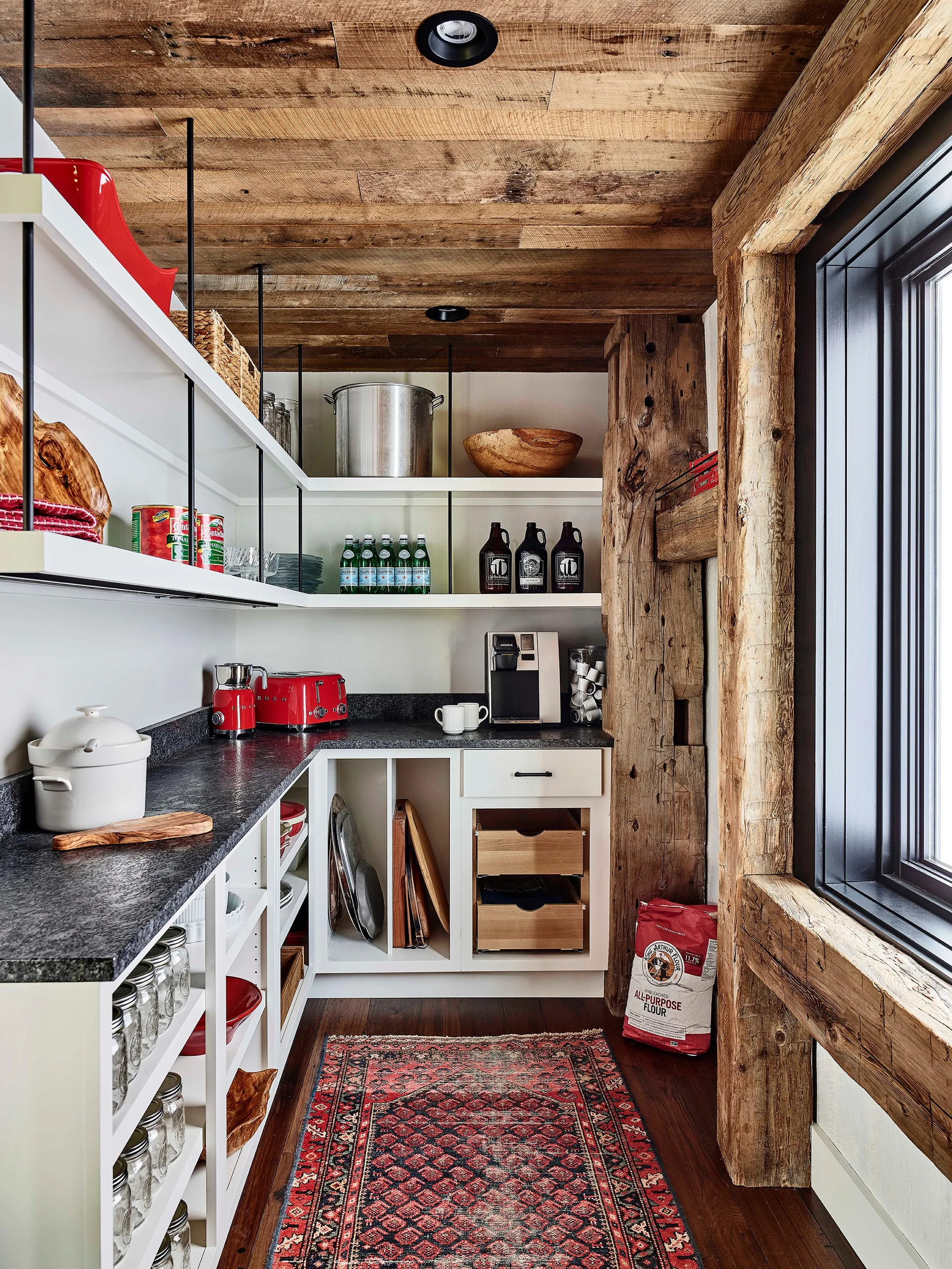 A rustic pantry features open white shelving, dark countertops, a reclaimed timber ceiling and post, and a red patterned runner rug.