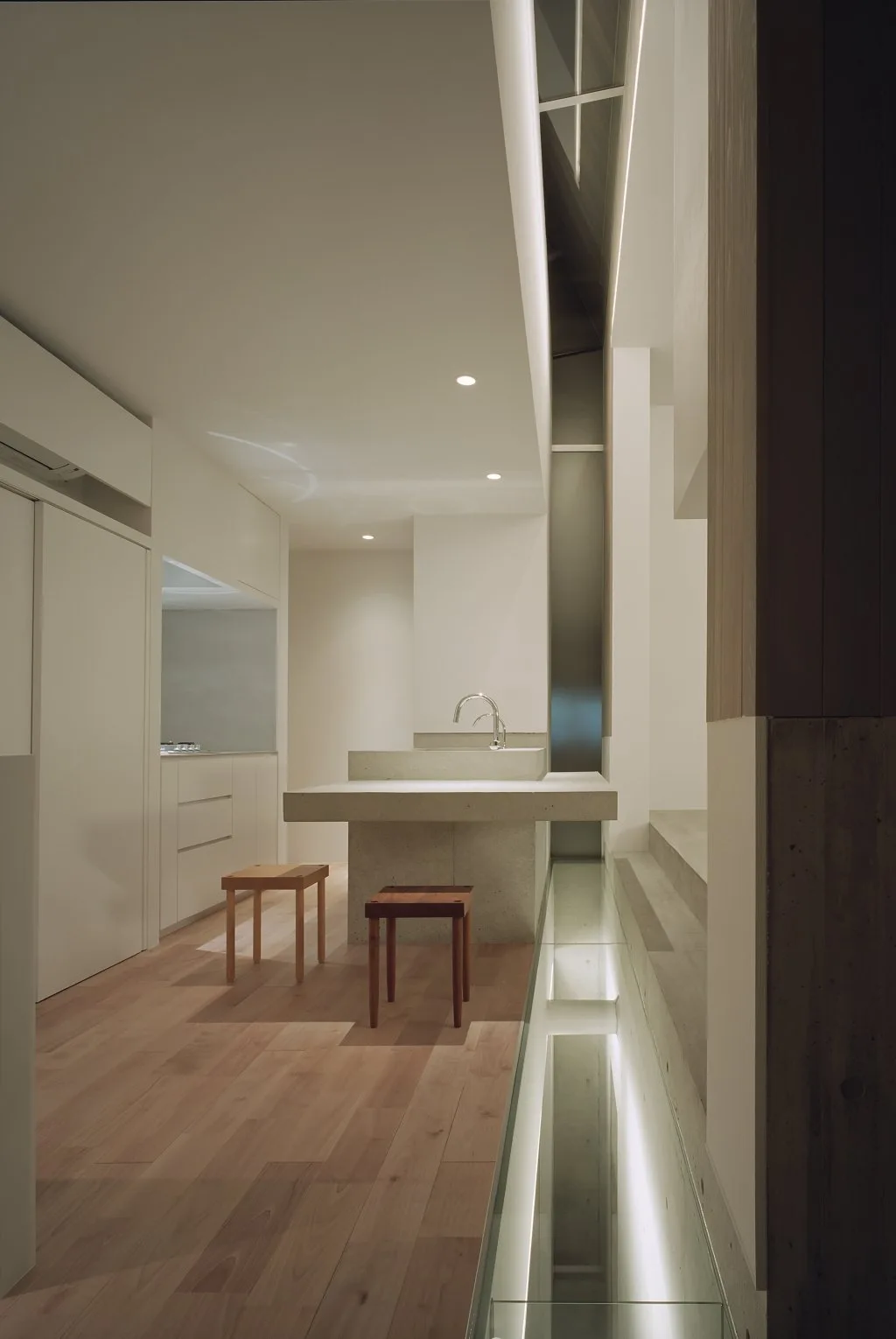 A minimalist kitchen features a cantilevered concrete island with wooden stools, flat-panel white cabinets, and lighting along concrete stairs.