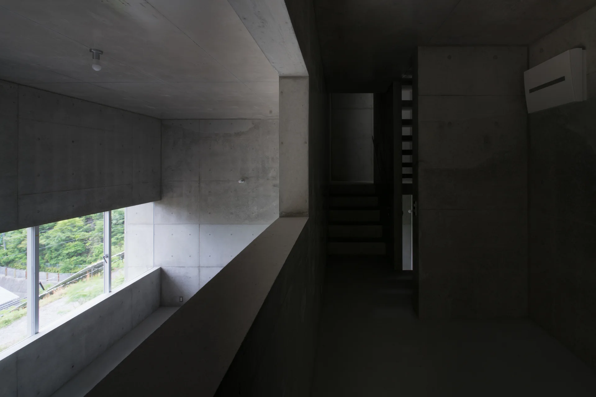 A minimalist raw concrete hallway with a viewing cutout overlooking a green hillside, featuring a staircase in the dark corridor.