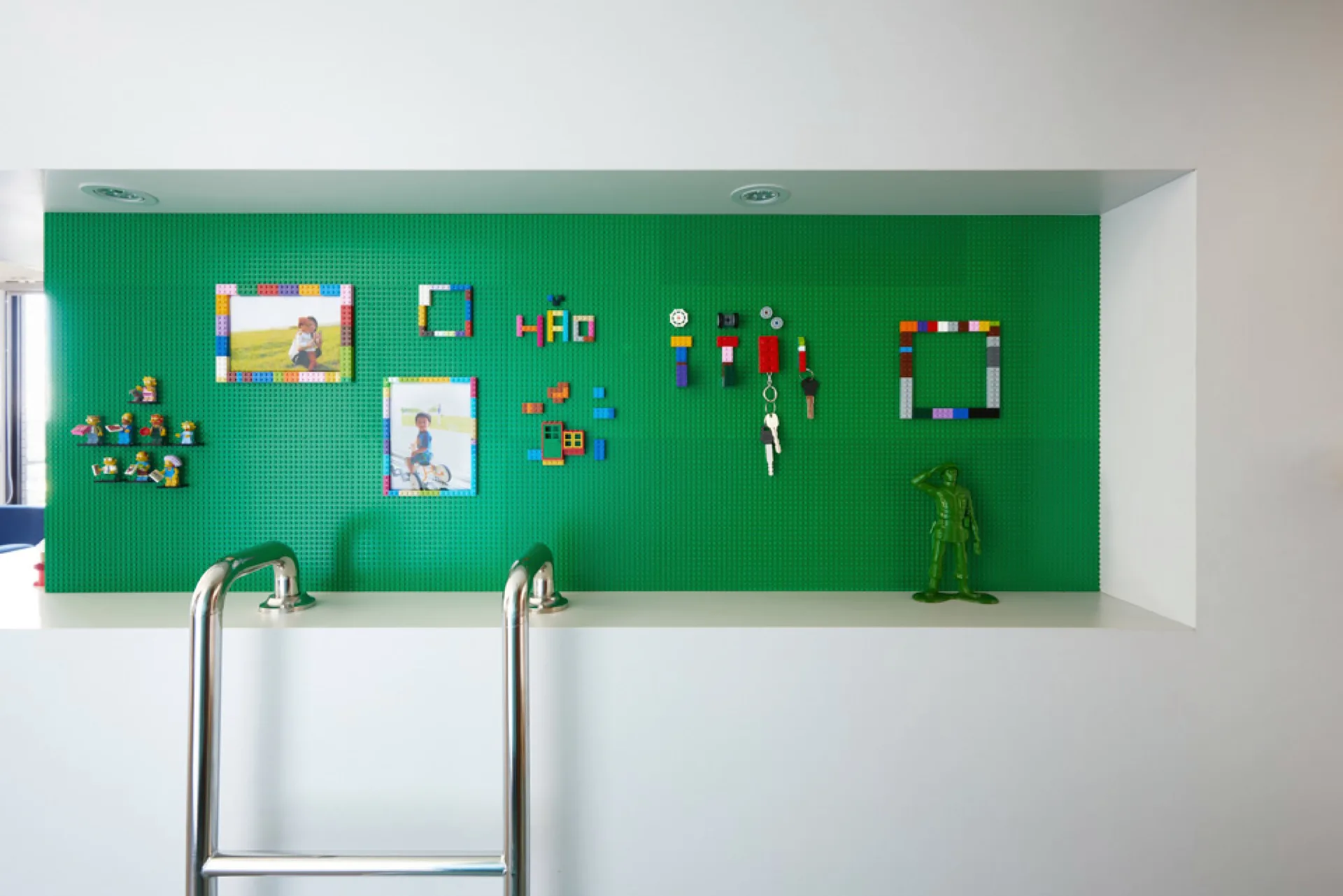 A recessed white wall niche lined with green Lego baseplates displaying photos in Lego frames, keys, and small toy figurines.