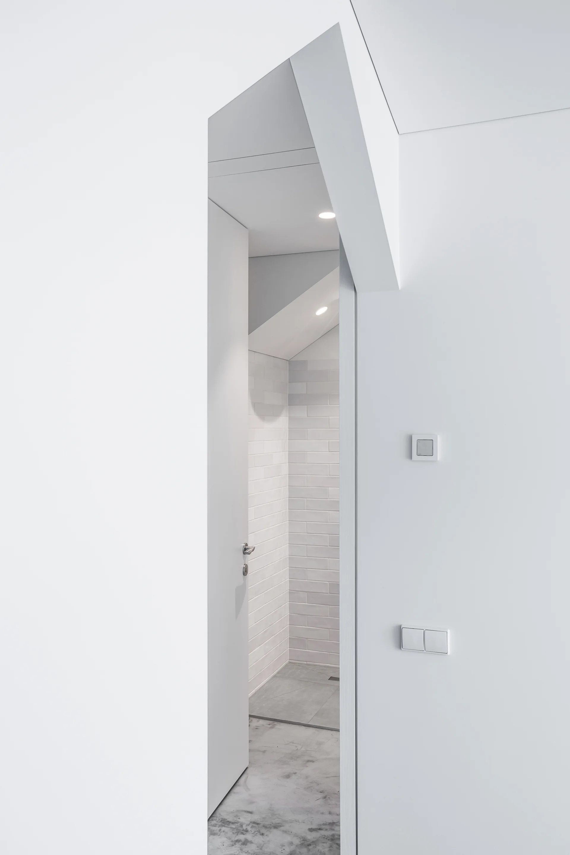 A view through a minimalist white doorway reveals a bathroom with light grey subway tile walls, a concrete floor, and recessed ceiling lights.