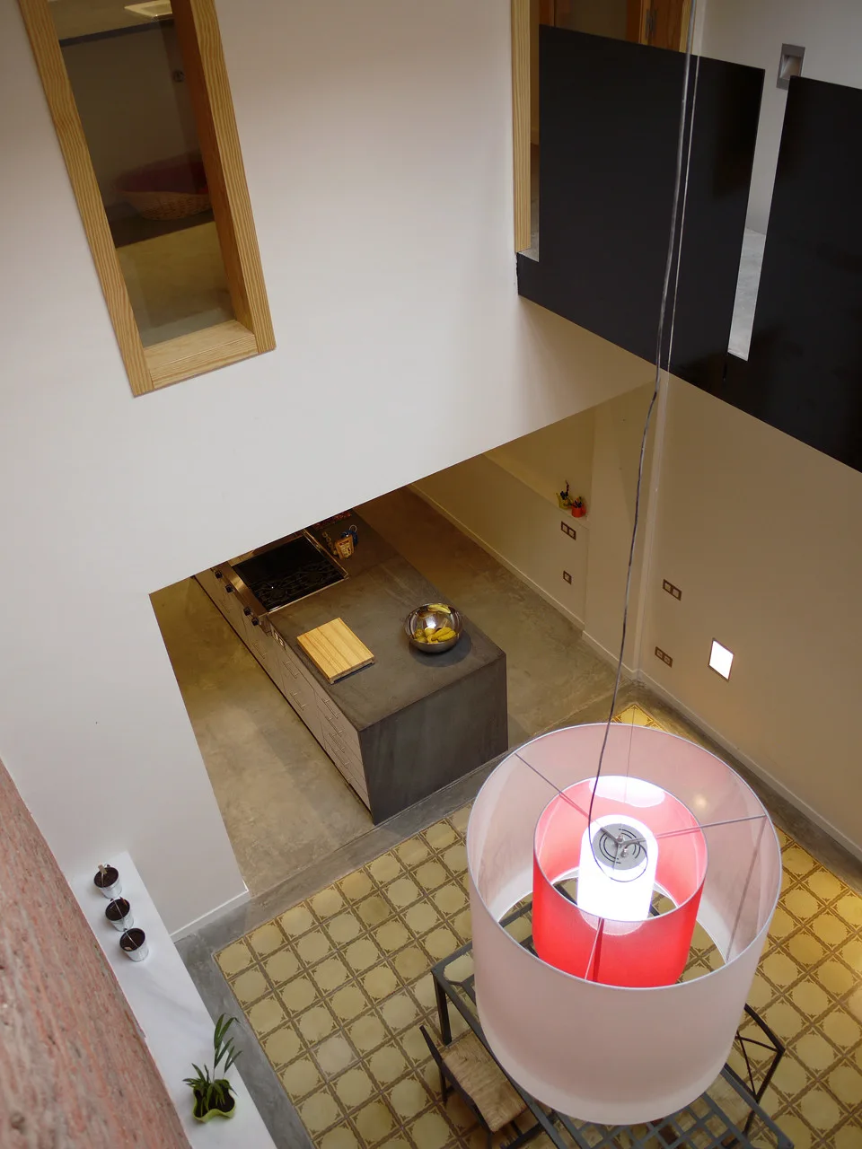 A high-angle view looking down at a concrete kitchen island, a large red and white pendant light, and geometric yellow tile flooring.