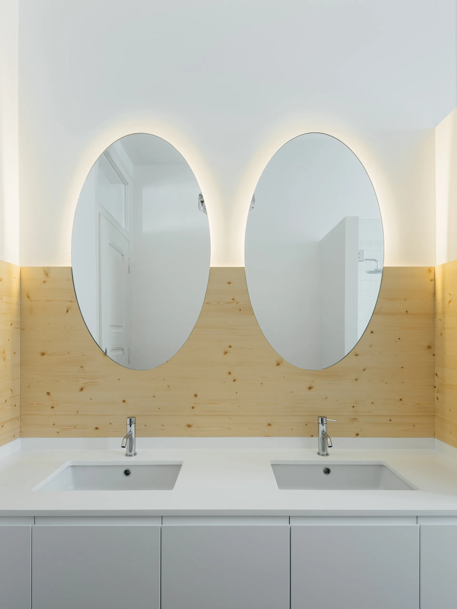 A modern double bathroom vanity with white countertop, two integrated sinks, chrome faucets, and backlit oval mirrors against a knotty wood paneled wall.