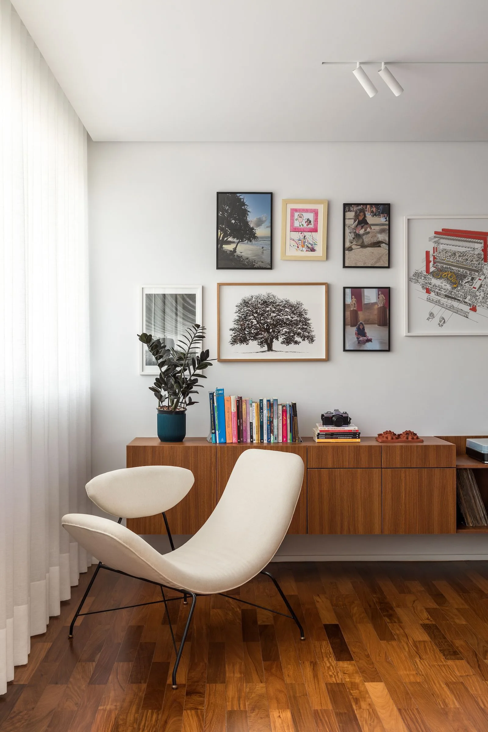 A white modern armchair stands on hardwood flooring next to a floating wooden sideboard beneath a curated gallery wall.