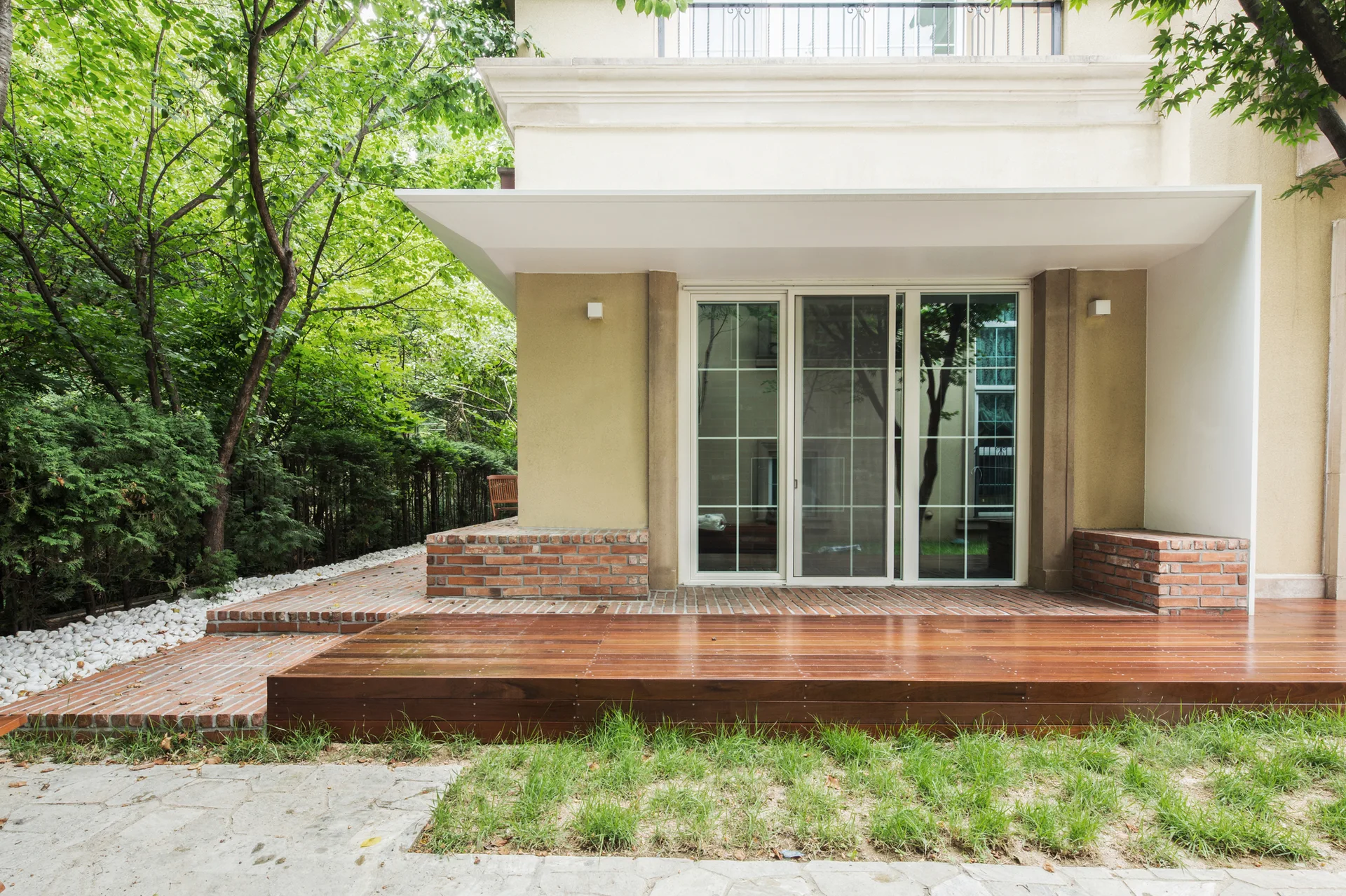 An outdoor patio features a raised wooden deck platform alongside red brick paving and sliding glass doors under a white overhang canopy.