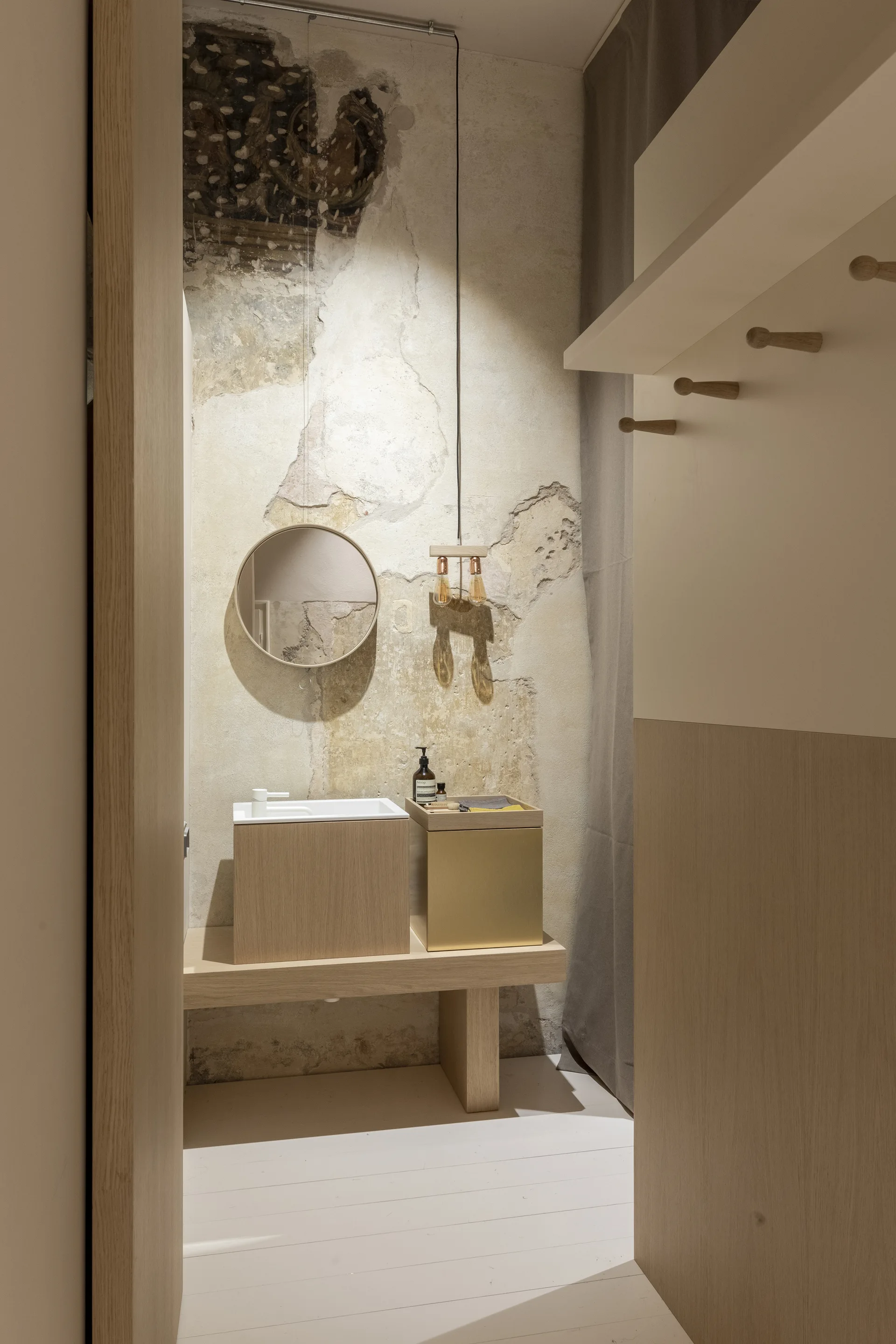 A modern bathroom vanity made of light wood and brass sits against a distressed plaster wall beneath a round mirror and a hanging bulb light.