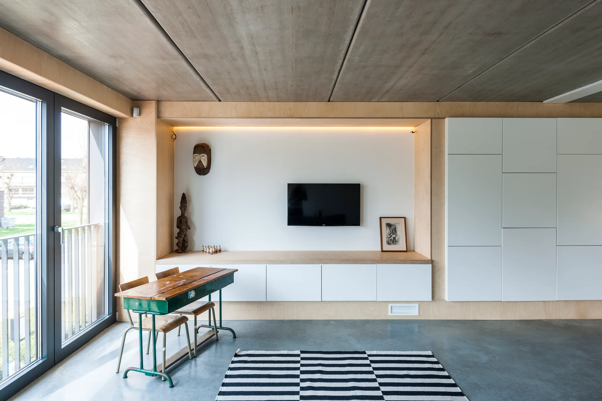 A minimalist room features an exposed concrete ceiling, plywood built-in cabinetry surrounding a TV, and a vintage wooden school desk near a large window.