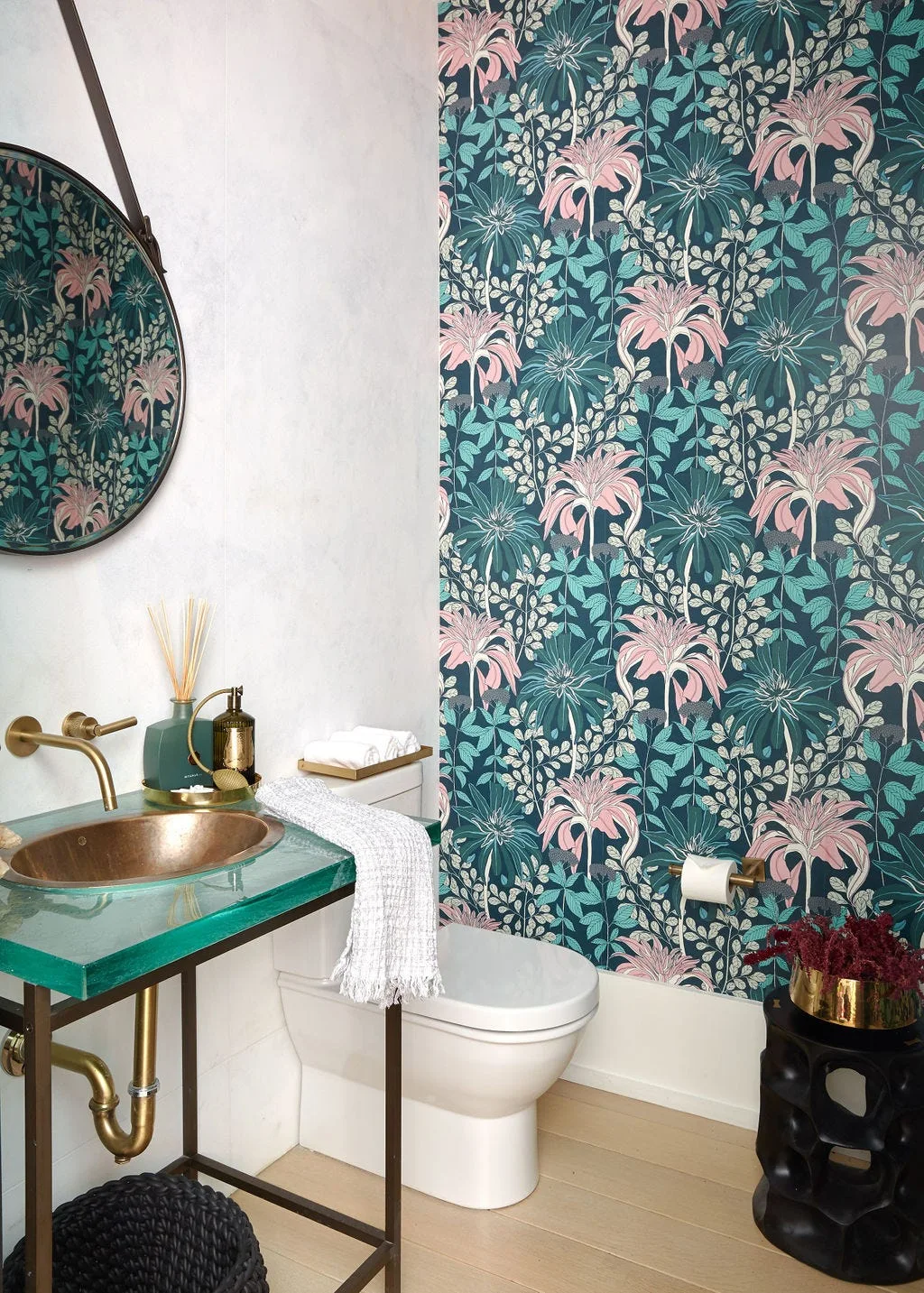 A powder room featuring bold teal floral wallpaper behind a toilet, alongside a green glass vanity with an integrated copper sink.