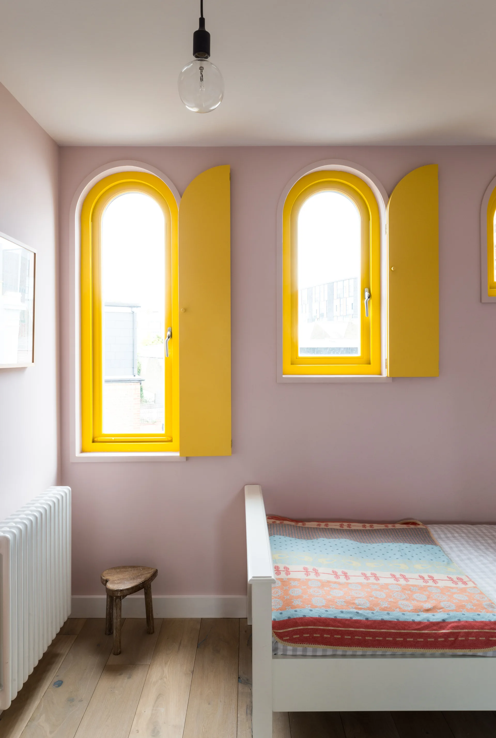 A pink bedroom with wooden flooring, a white bed frame, a rustic stool, and two arched windows featuring bright yellow painted frames and shutters.