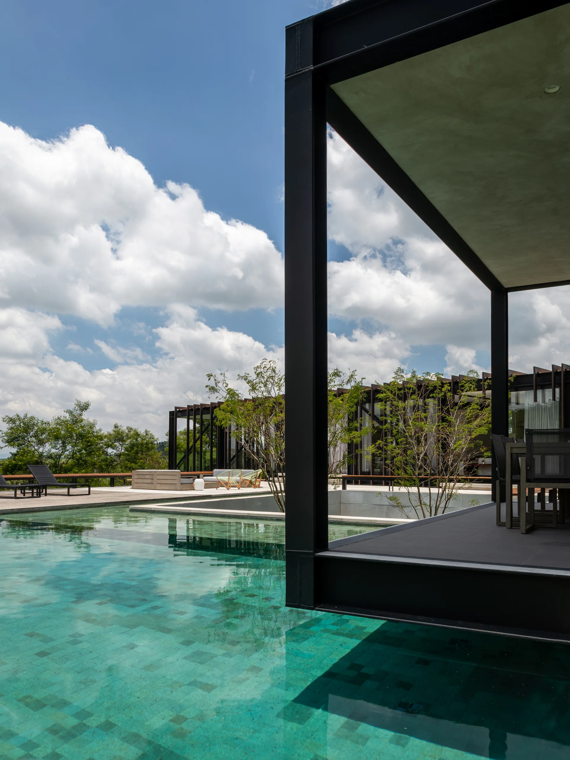 A modern turquoise swimming pool sits next to a wooden deck with a black steel frame pavilion, lounge chairs, and green trees under a blue sky.
