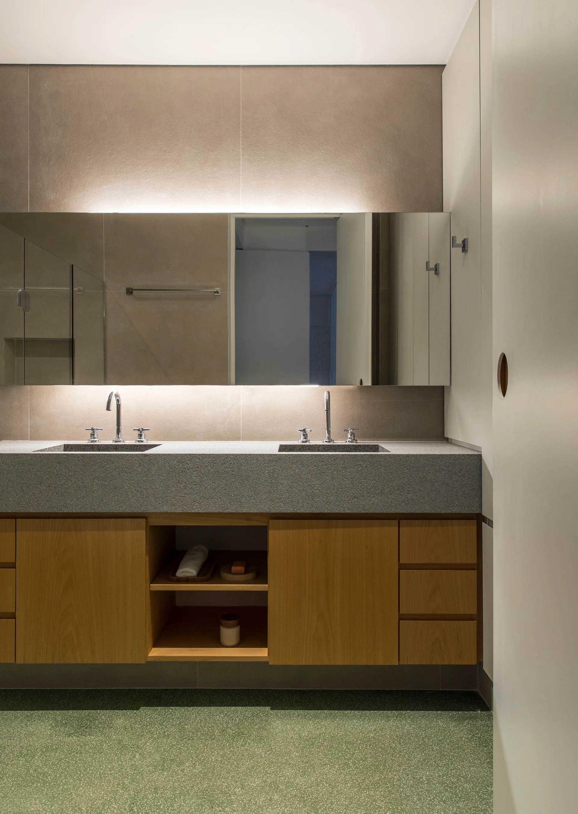 A wood bathroom vanity with a thick grey countertop and double sinks sits below a large backlit mirror, above green speckled terrazzo flooring.