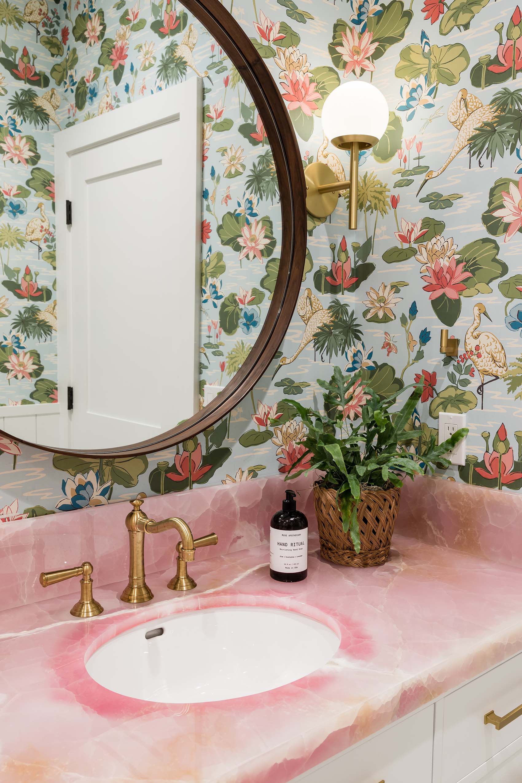 A powder room features a pink stone vanity with a brass faucet, a round wood-framed mirror, and blue botanical wallpaper with bird motifs.