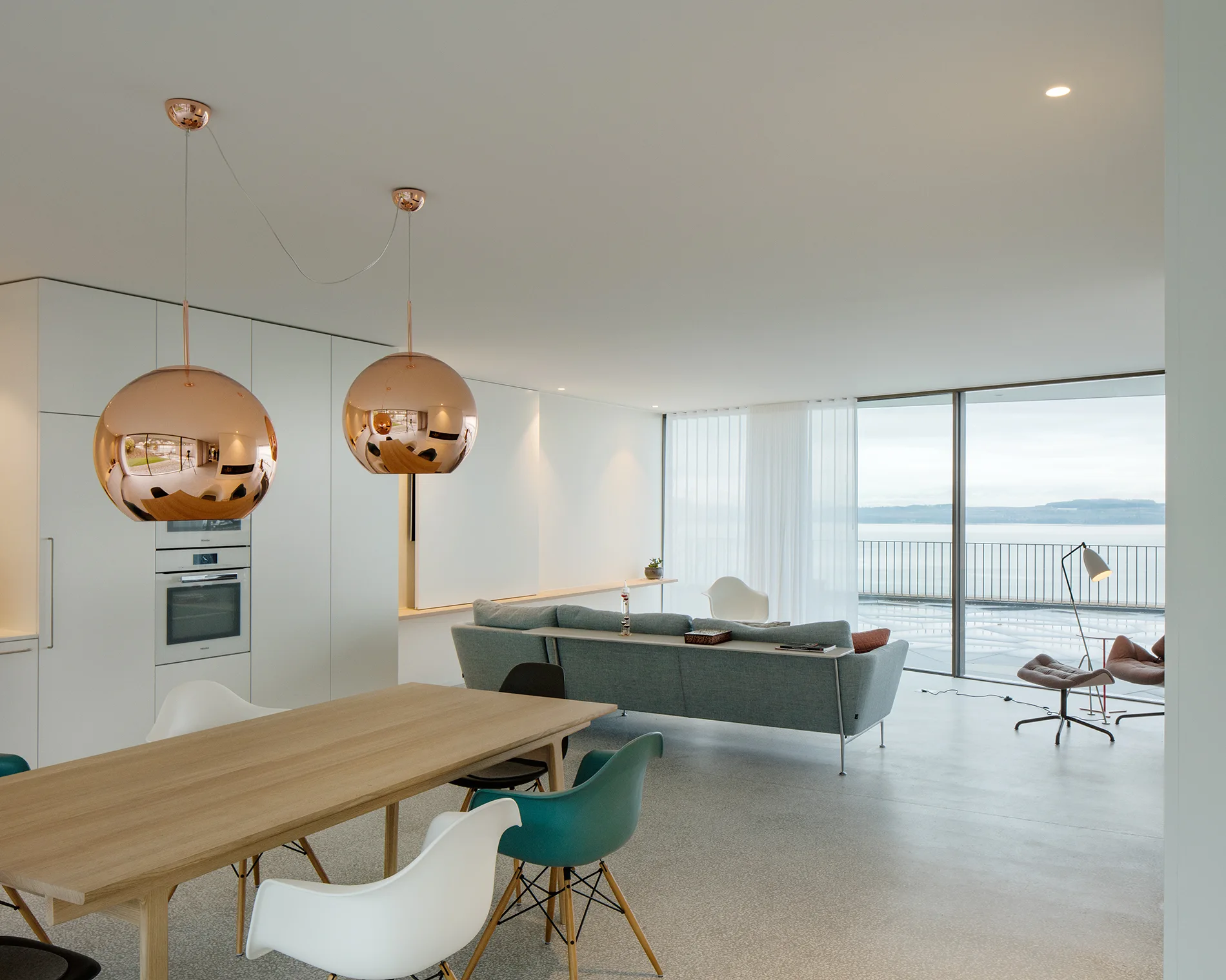 An open-plan room features a wooden dining table under two copper globe pendants, a light blue sofa, and large windows with water views.