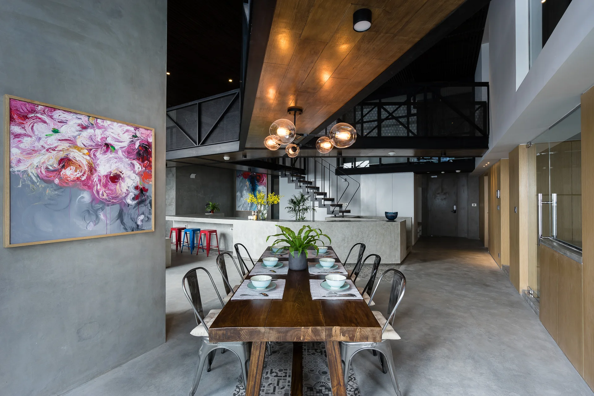 An industrial dining room features a wooden table, metal chairs, concrete walls, a globe pendant light, and a colorful floral painting.