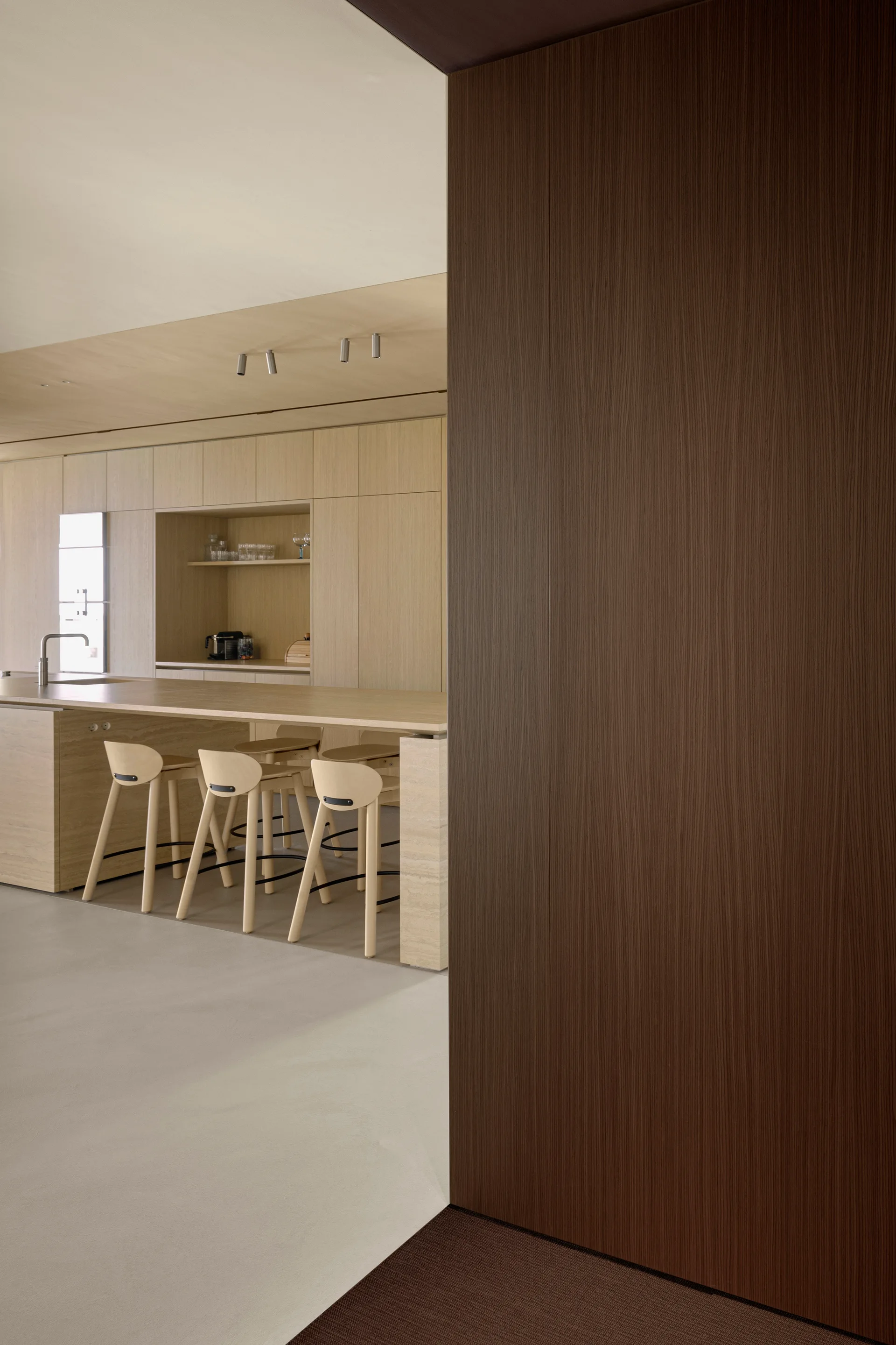 A minimalist kitchen featuring seamless light wood cabinetry, a central island with wood bar stools, and a dark wood partition wall in the foreground.