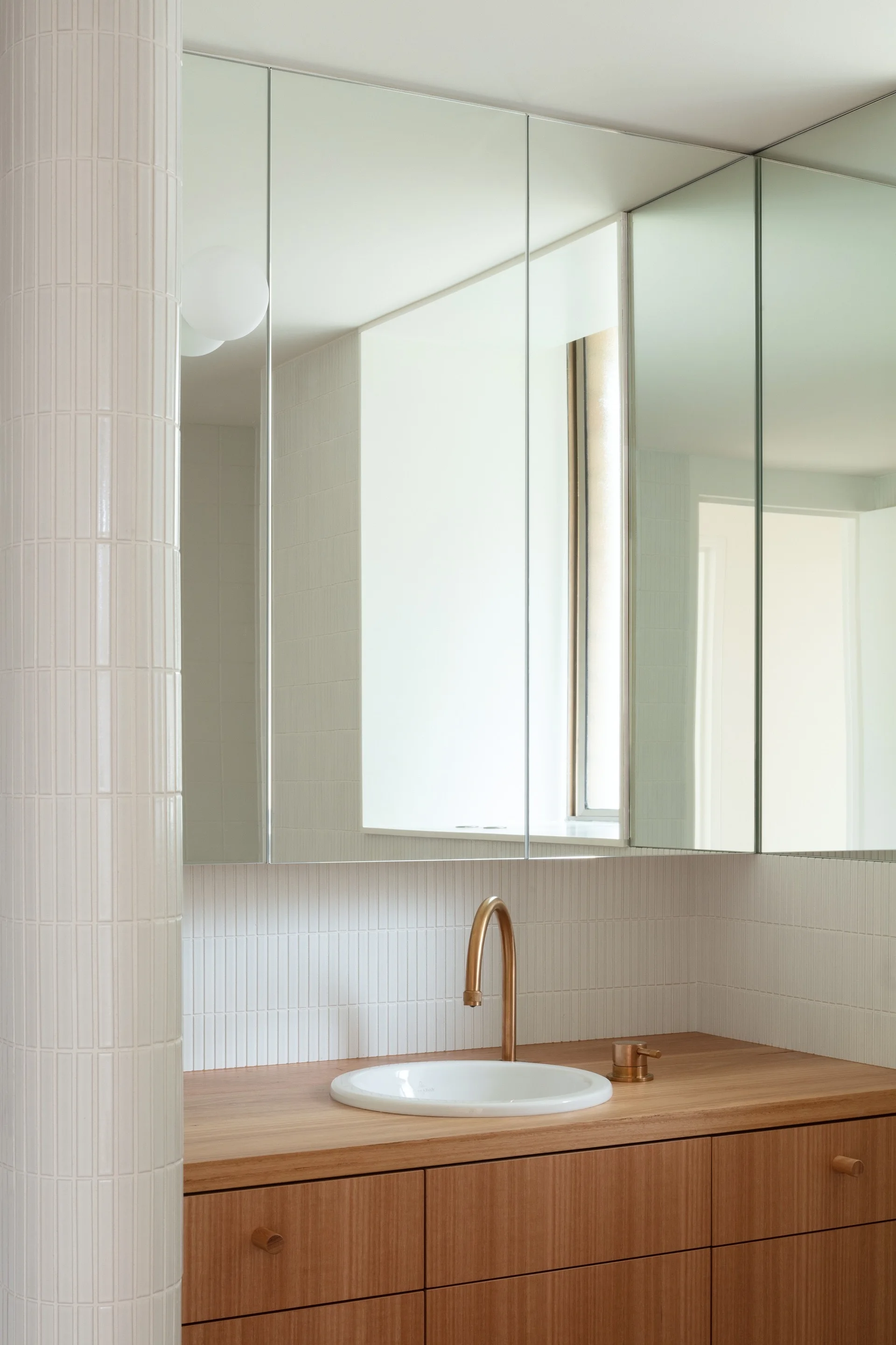 A modern bathroom corner features a wood vanity with a white basin, a brass faucet, a vertical white tile backsplash, and a tiled column.