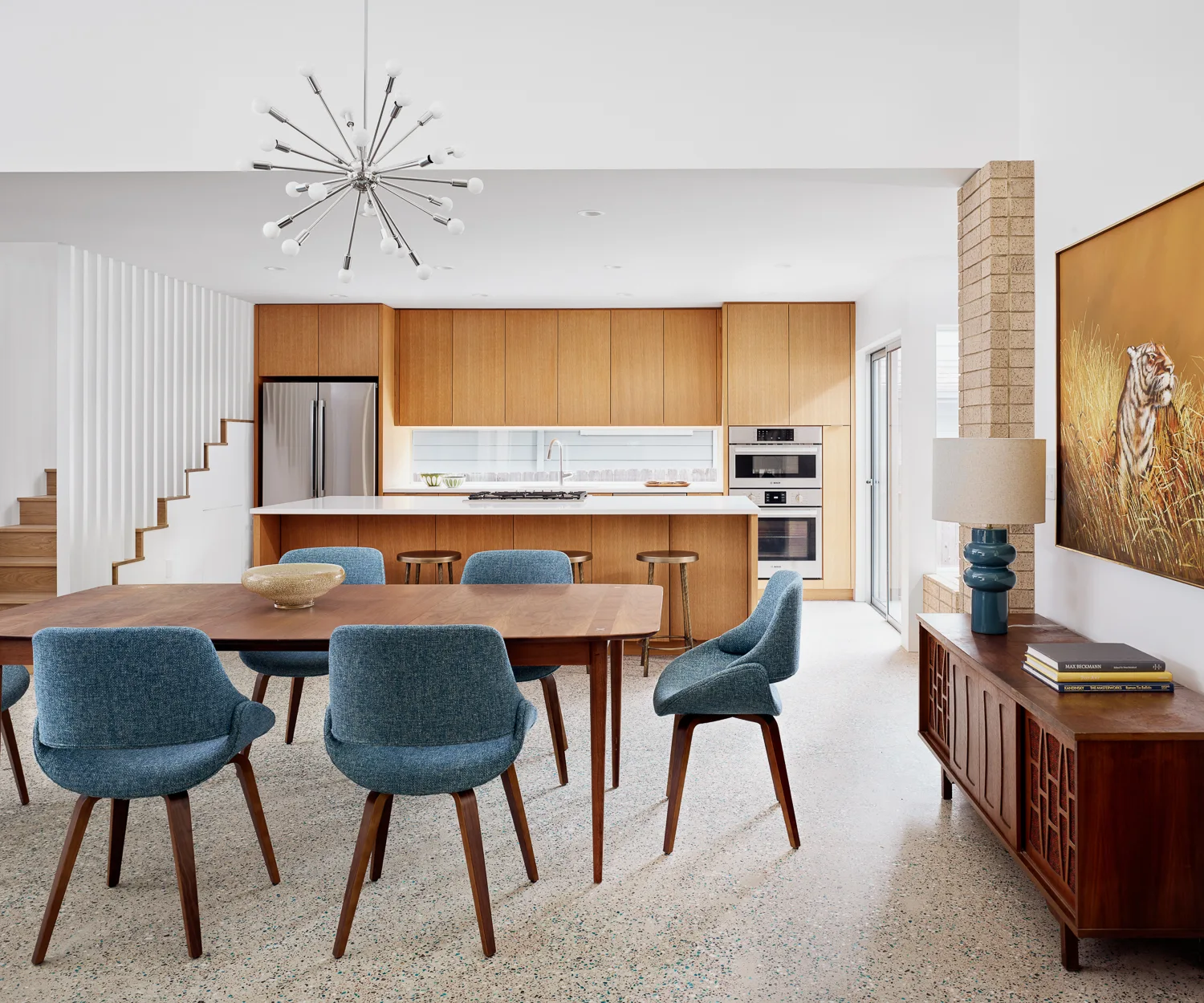 A mid-century modern open-plan kitchen and dining area features natural wood cabinetry, blue upholstered chairs around a wooden table, and terrazzo flooring.