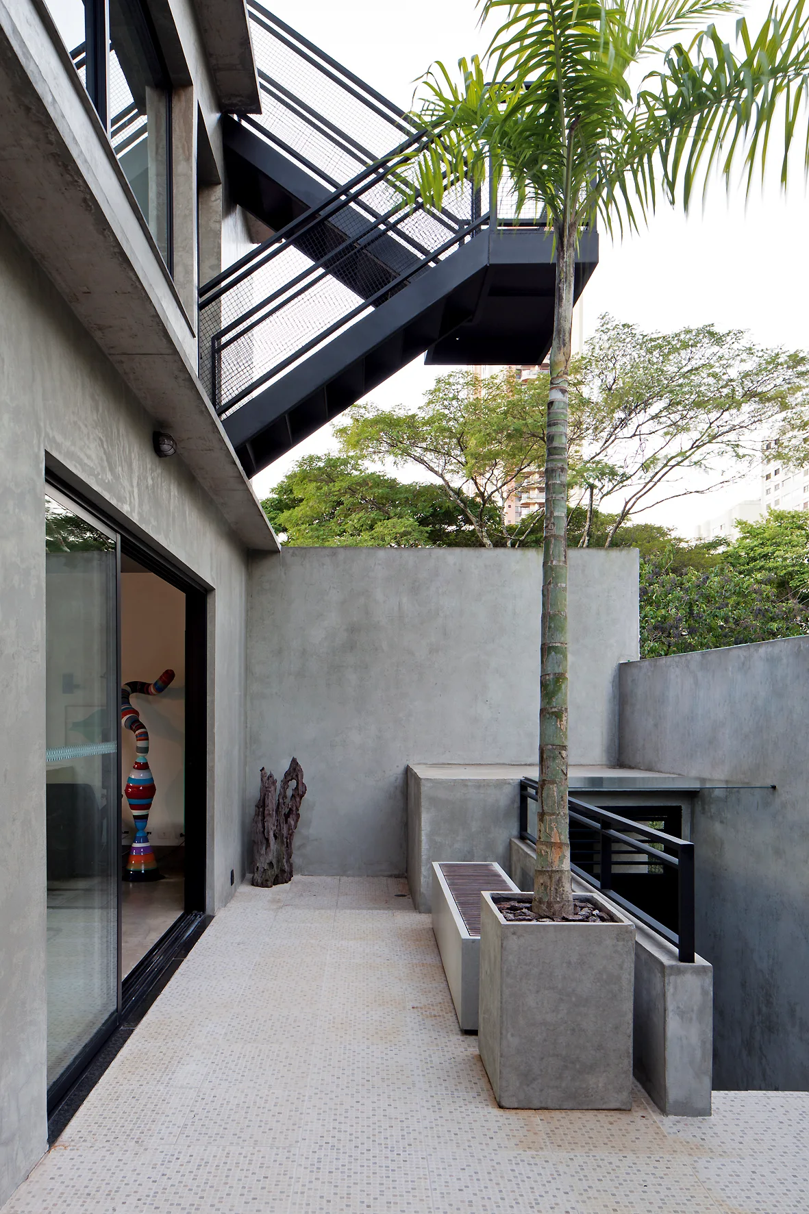 A modern concrete terrace features a black industrial metal staircase, a built-in bench, and a tall palm tree planted in a square concrete planter.
