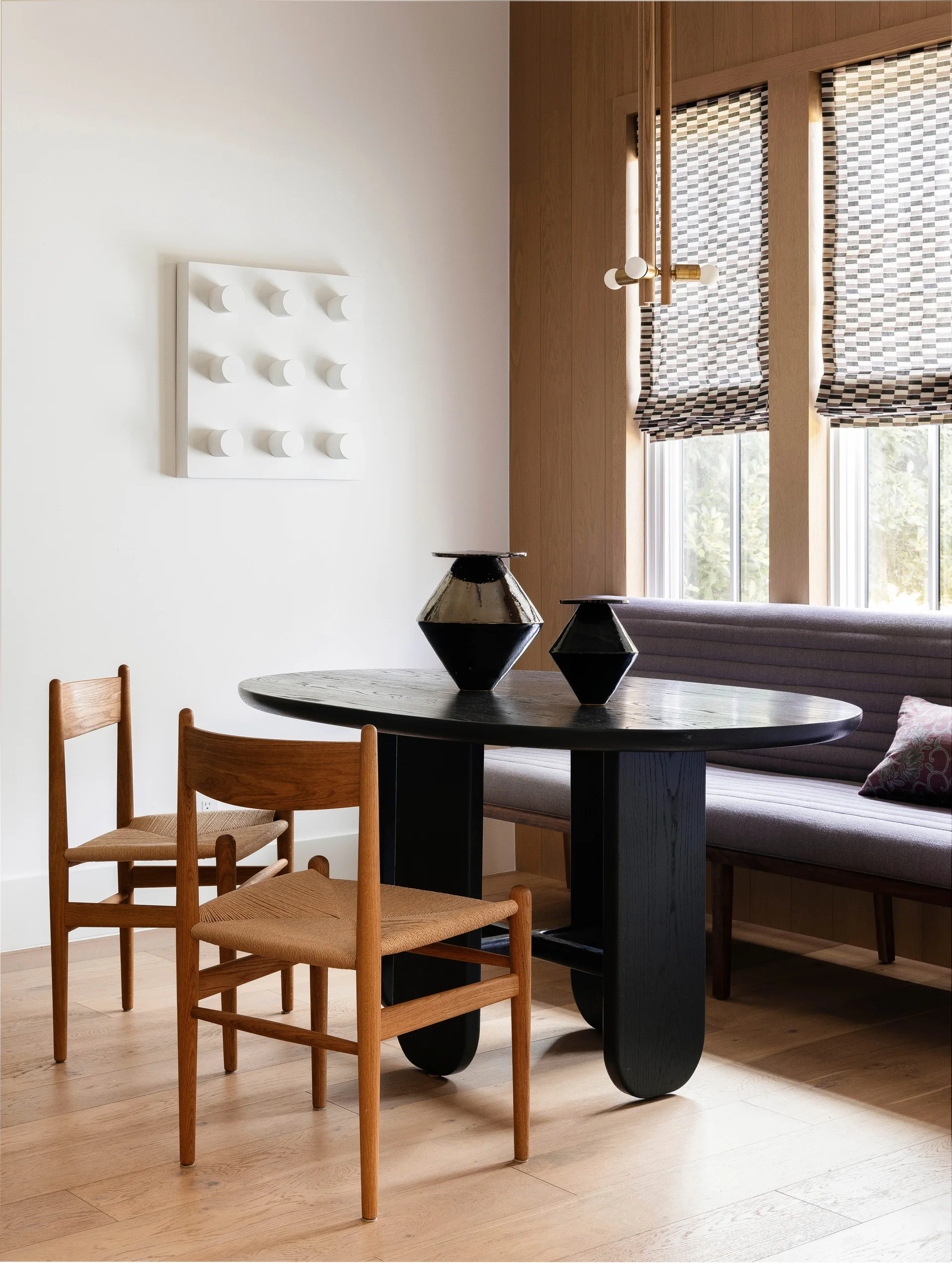 A modern dining area features a black oval table, wooden chairs with woven seats, and a gray banquette against wood-paneled walls.