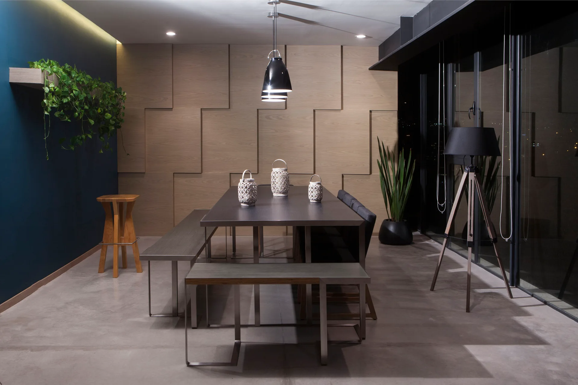 A modern dining room features a dark table with benches set against a geometric wood-paneled accent wall, a blue side wall, and a black pendant light.