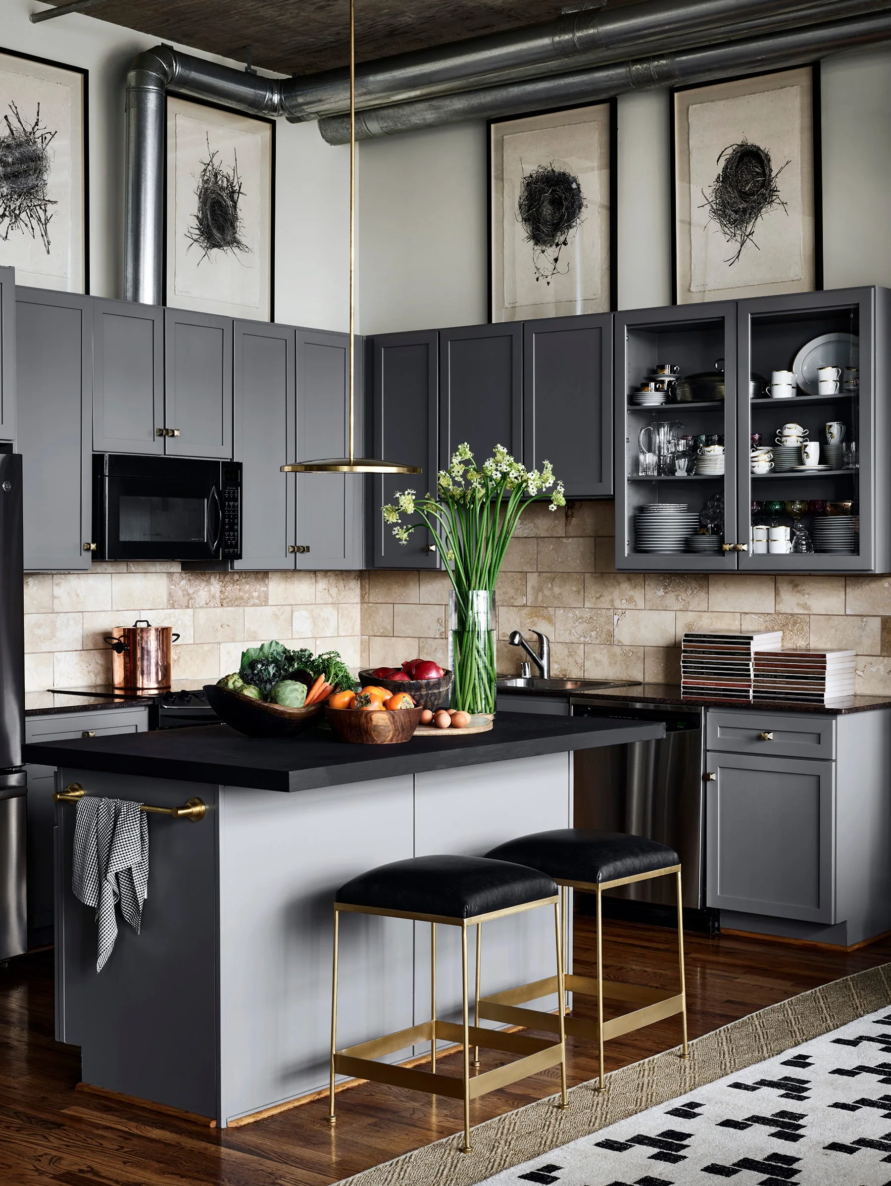 A modern kitchen featuring gray cabinetry, a black-topped island with two brass stools, a stone tile backsplash, and exposed ceiling ductwork.